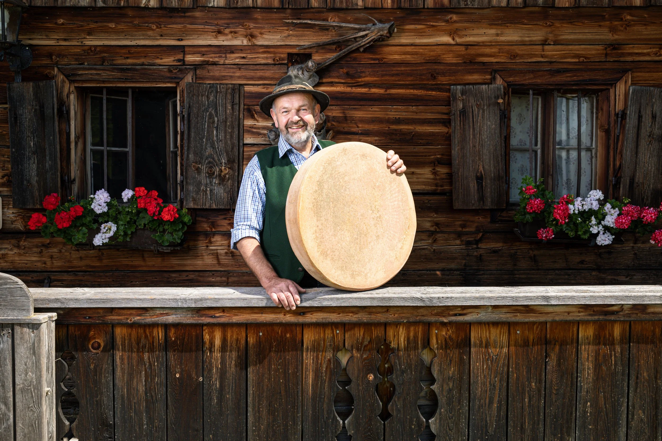 Ein lachender Mann in traditioneller Kleidung mit Hut steht vor einer Holzwand und hält einen großen Käse. Im Hintergrund sind zwei Fenster mit Blumenkästen voller bunter Blumen, und es sind Tierköpfe und ein Gewehr an der Wand zu sehen.