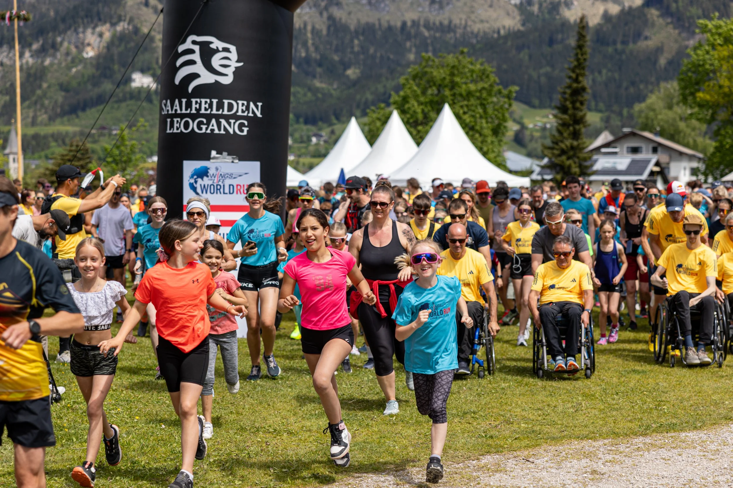 Gruppen von Menschen, darunter Kinder, laufen in einem Park bei einem Event inmitten grüner Landschaft mit Baumkronen und Hügeln im Hintergrund. Es ist sonnig und festlich, mit großen weißen Zelte im Hintergrund.