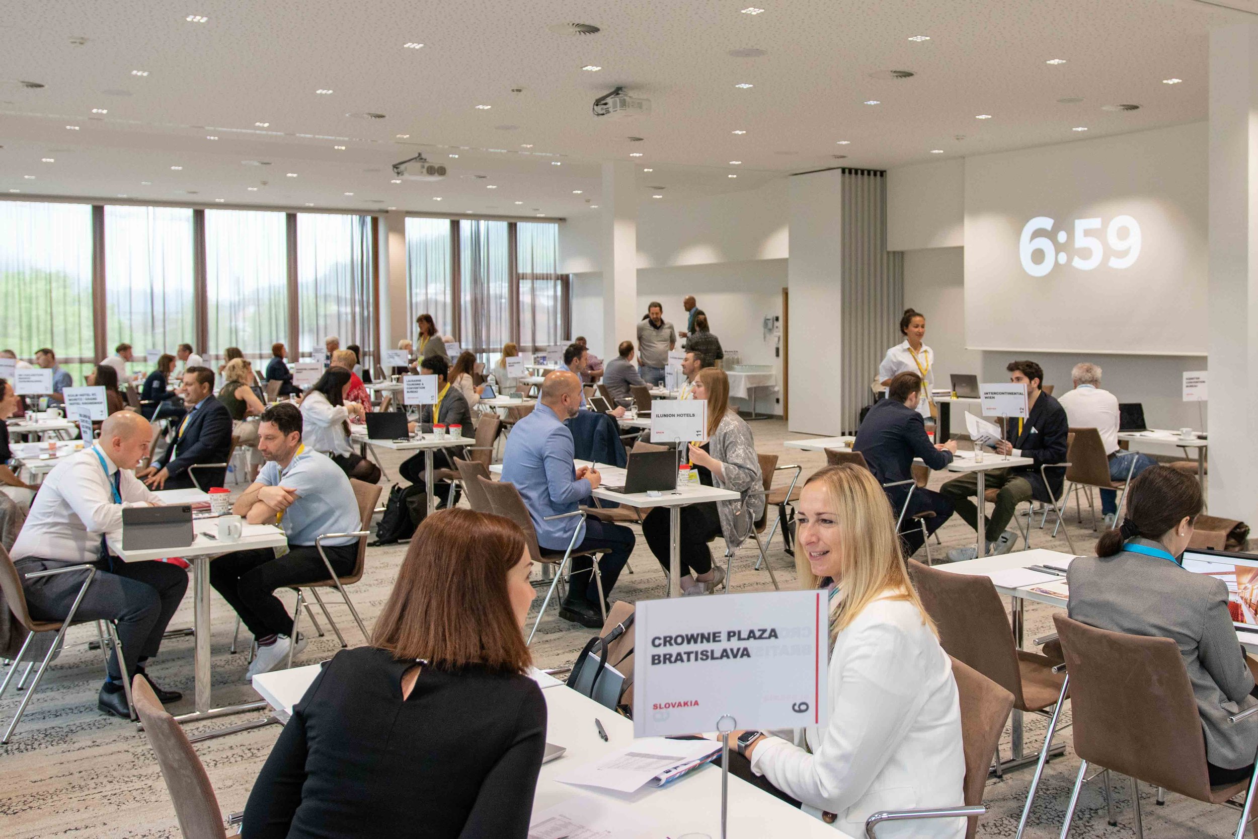 Geschäftsleute in einem großen, hellen Konferenzraum, die an Tischen sitzen, reden und Laptops verwenden, mit einem großen Bildschirm, auf dem die Uhrzeit 6:59 angezeigt wird.