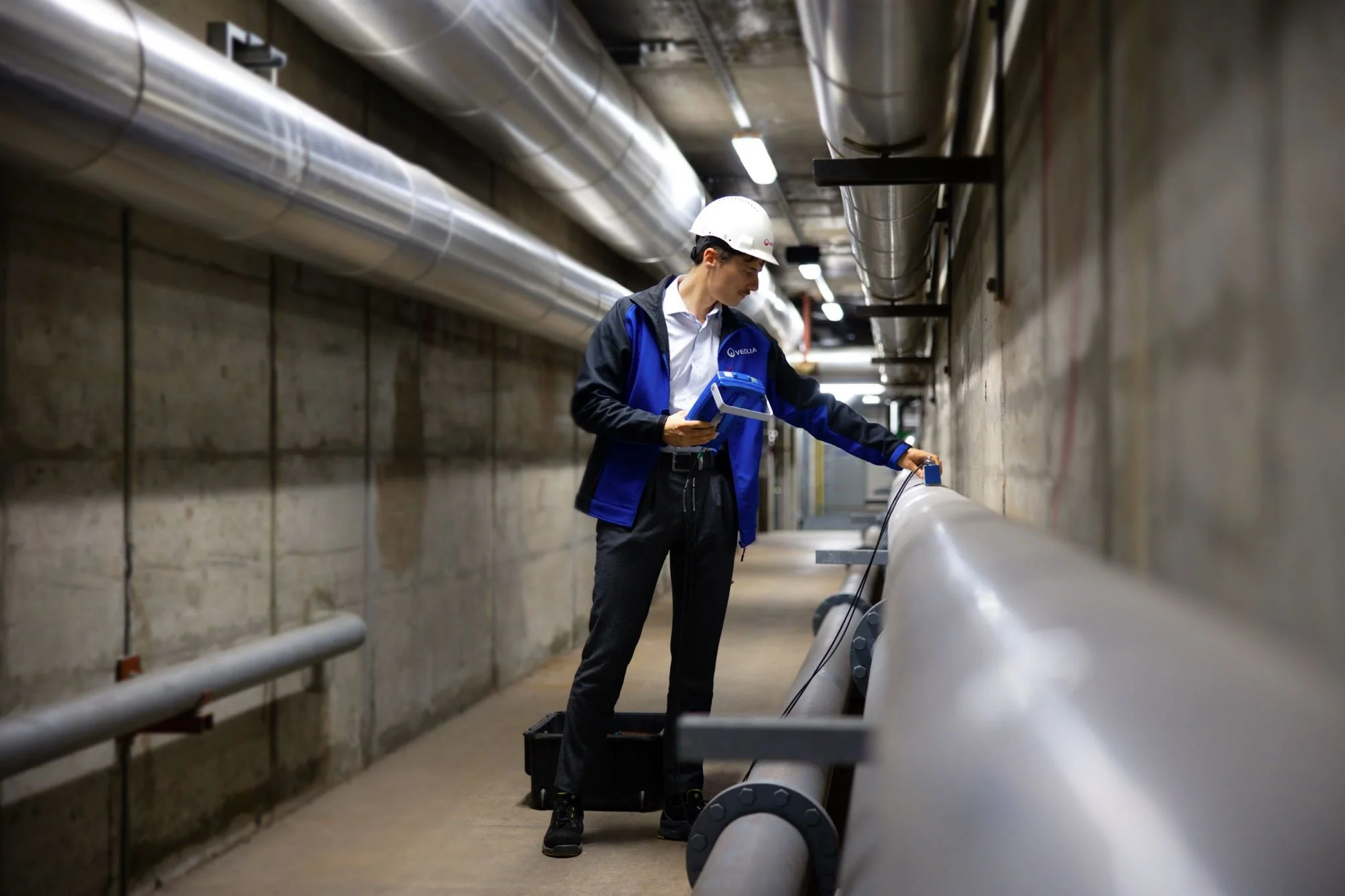 Mann in Sicherheitshelm und blauer Jacke überprüft eine große Rohrleitung in einem technischen Kellerraum.