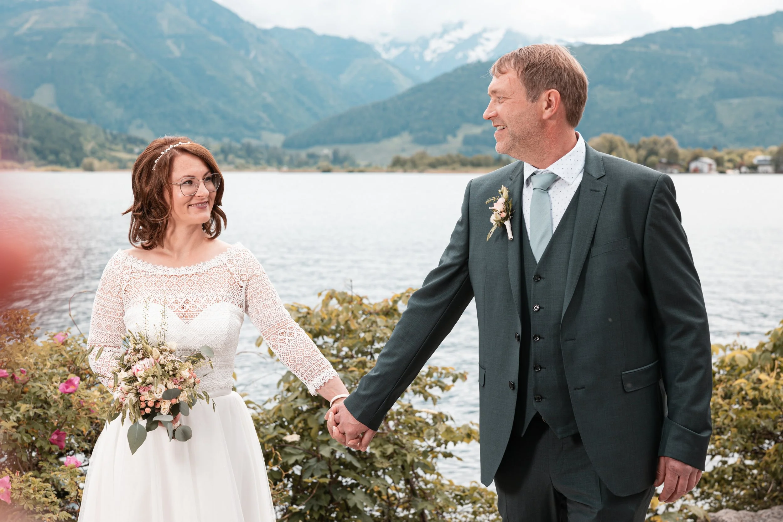 Ein Brautpaar bei einer Hochzeit am See, umgeben von Bergen, die Hand in Hand halten.