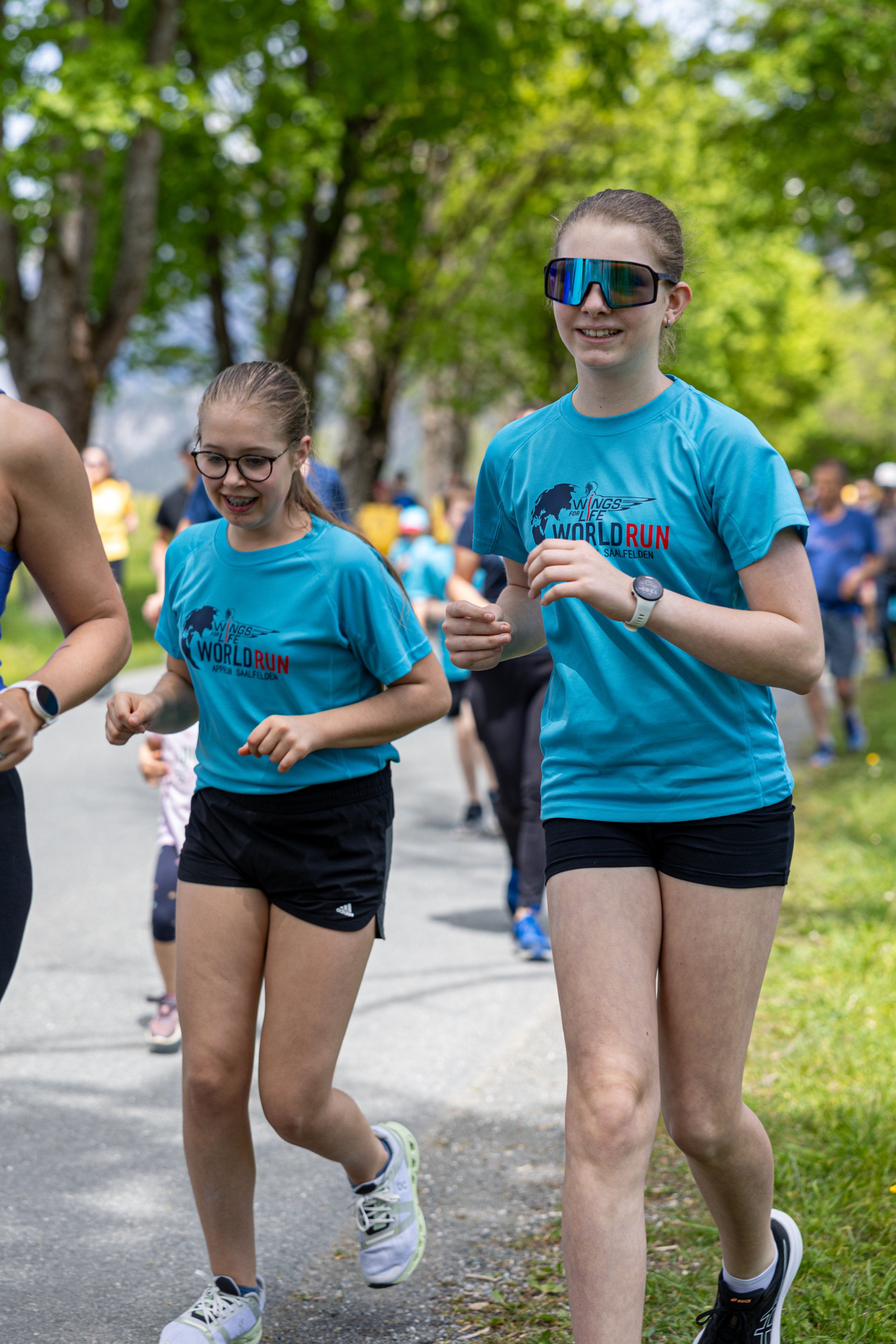 Zwei junge Läuferinnen bei einem Straßenlauf, beide tragen blaue T-Shirts mit dem Aufdruck "World Run" und laufen auf einem Weg in einem Park mit grünen Bäumen.