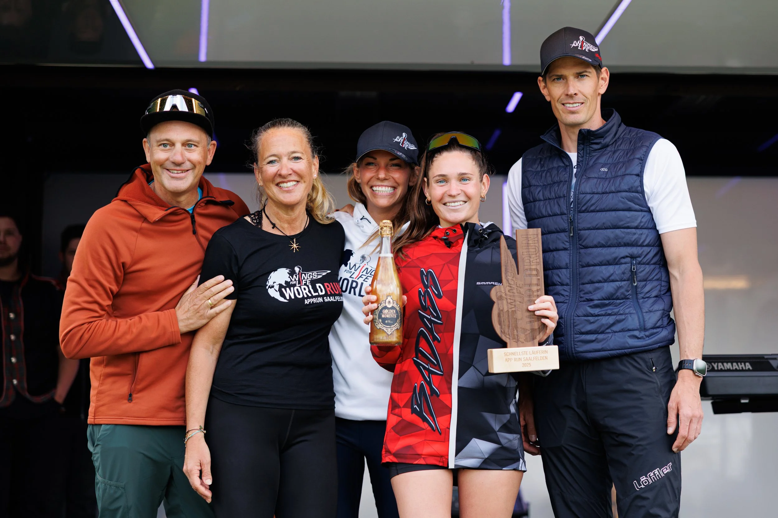 Gruppe von fünf Menschen, die bei einer Siegerfeier lachen und feiern. Eine Frau in sportlicher Kleidung hält eine Trophäe und eine Flasche Sekt. Es sind Männer und Frauen, einige tragen Sportkleidung und Mützen. Der Hintergrund ist dunkel mit Neonli