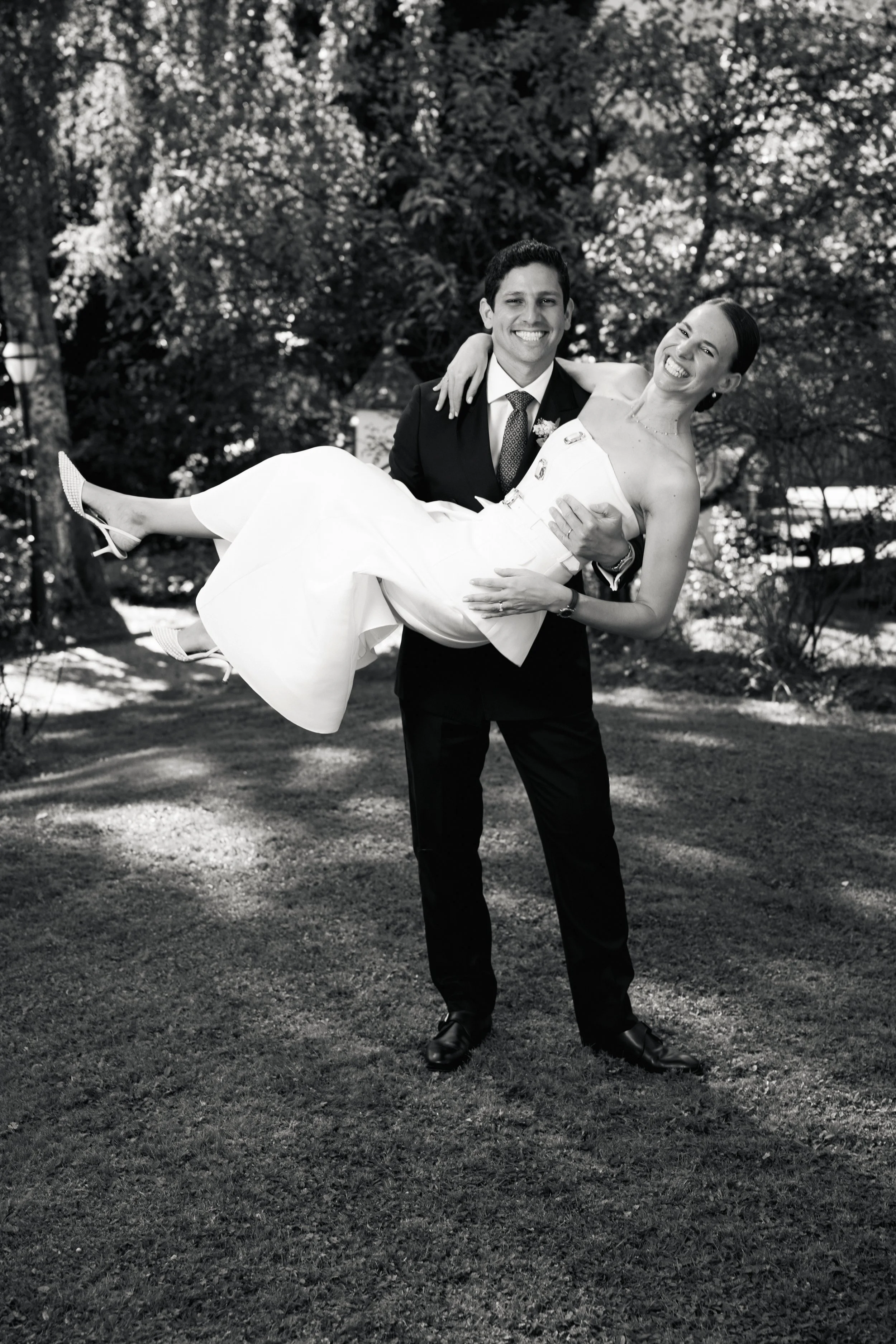 Ein Mann in einem Anzug hält eine lachende Frau in einem weißen Kleid in einem Park. Die Frau wird hochgehoben, während beide smiling sind.