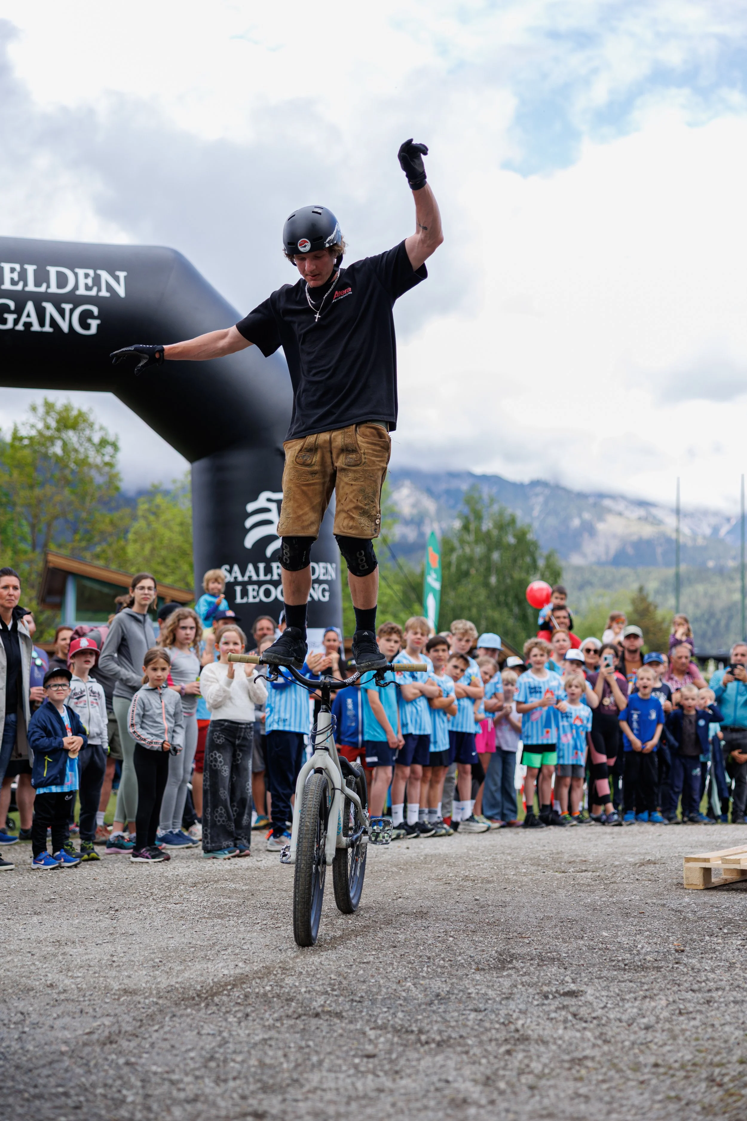 Mann macht Balanceakt auf Fahrrad vor Publikum bei einem Outdoor-Event, mit Berg im Hintergrund.