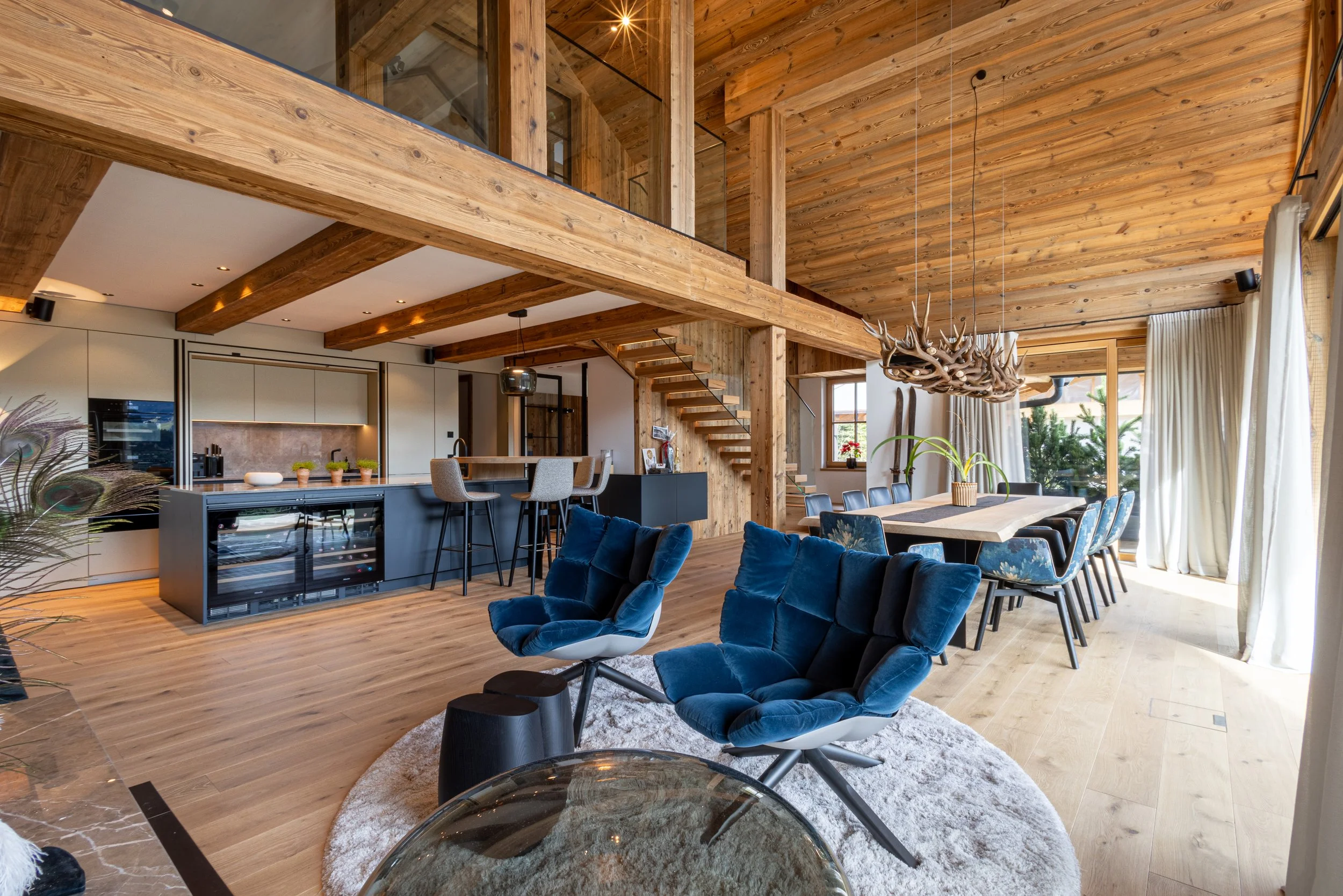 Offenes Wohnzimmer und Küche mit Holzdecke, Treppe im Hintergrund, Sesseln und Esstisch, viel Tageslicht durch große Fenster.