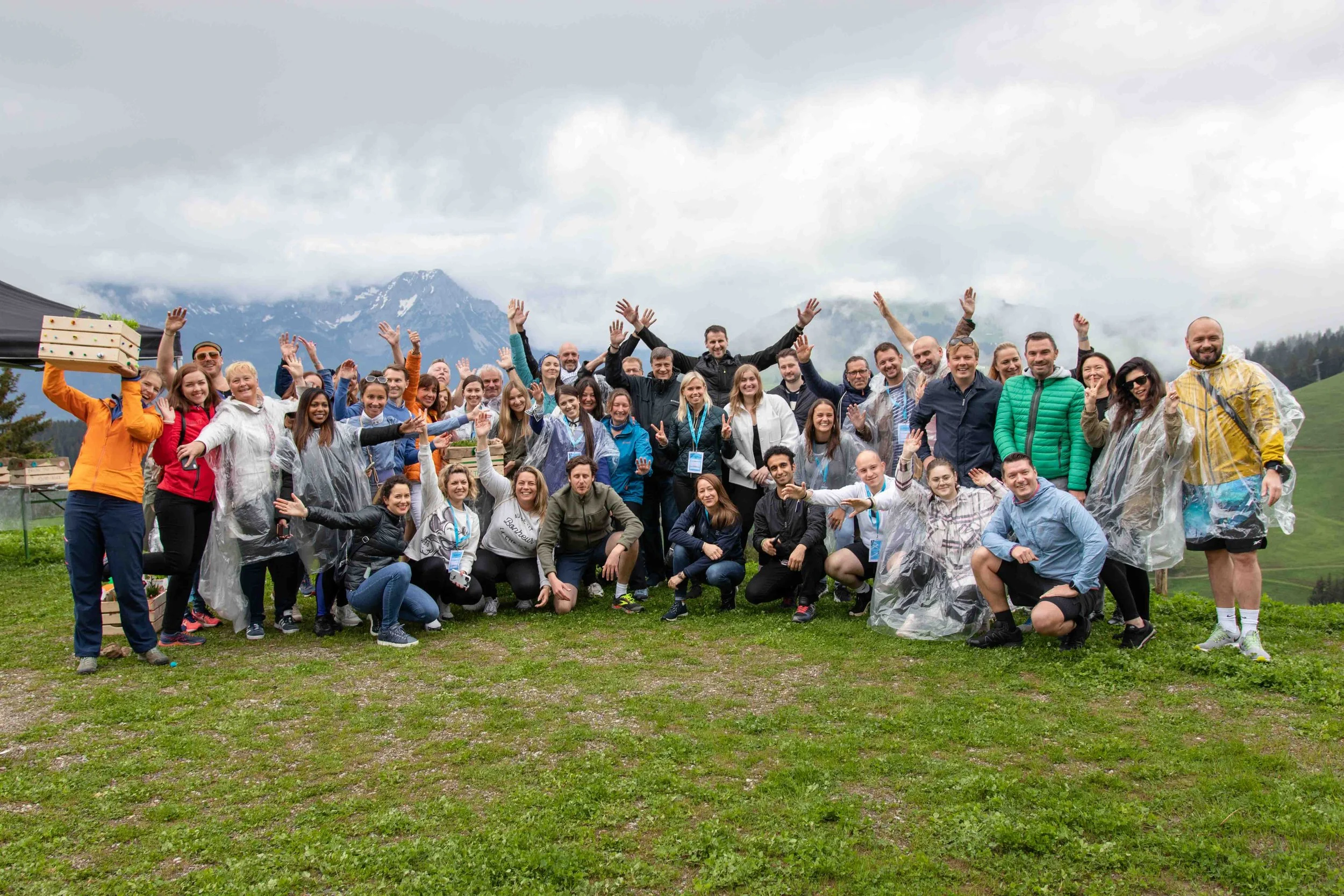 Eine große Gruppe von Menschen steht im Freien auf einer Wiese, umgeben von Bergen und bewölktem Himmel, alle lachen und winken zur Kamera.