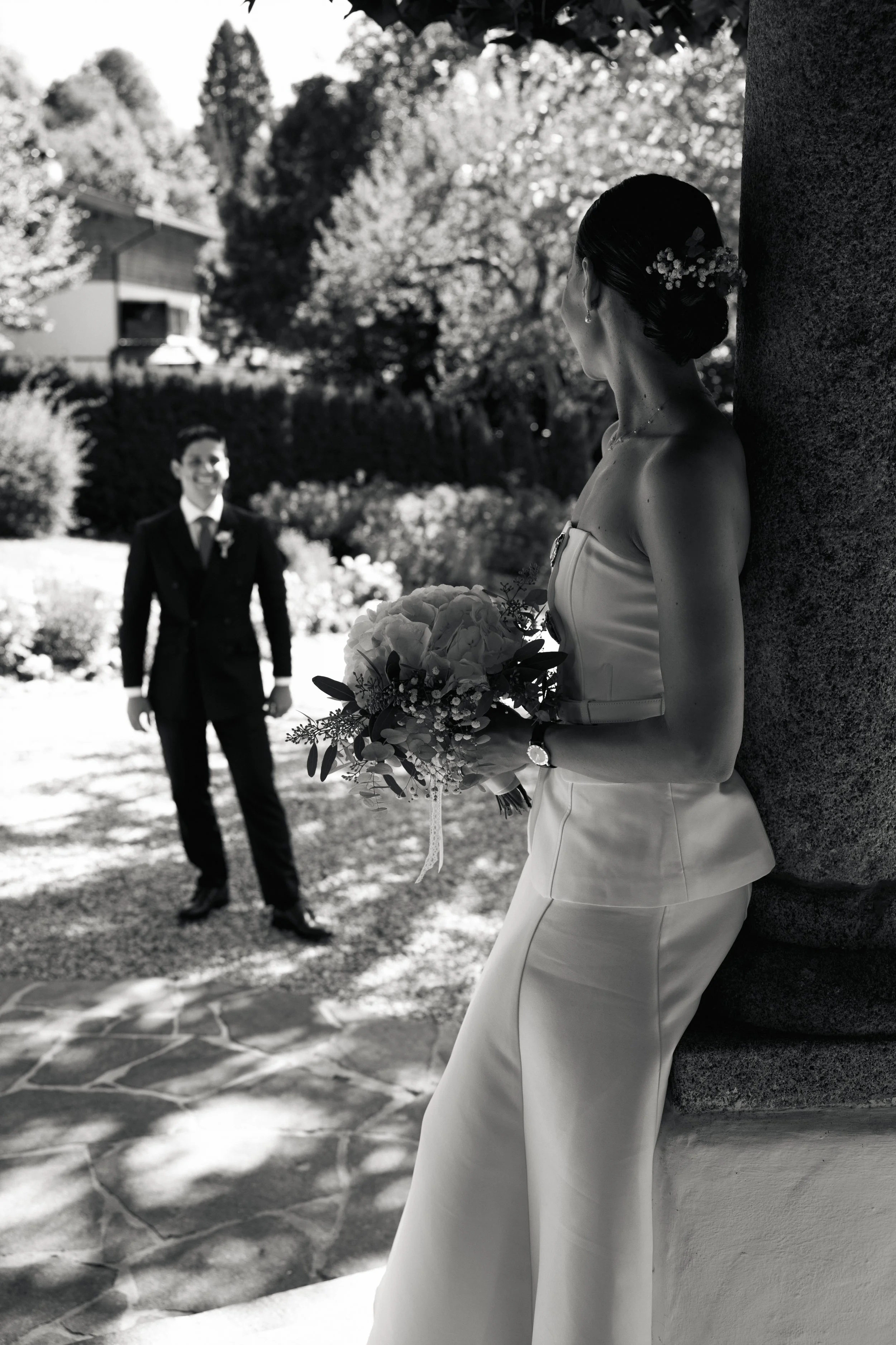 Braut mit Blumenstrauß lehnt an einer Säule, während der Bräutigam im Garten steht und auf sie schaut, bei Sonnenlicht an einem Hochzeitstag im Freien.