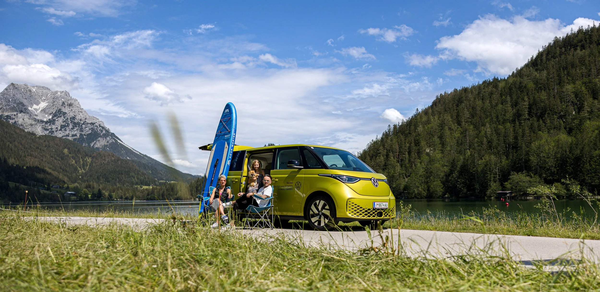 Familie mit zwei Kindern sitzt vor einem gelben Elektroauto am Fluss in den Bergen, mit Paddelboards im Hintergrund.