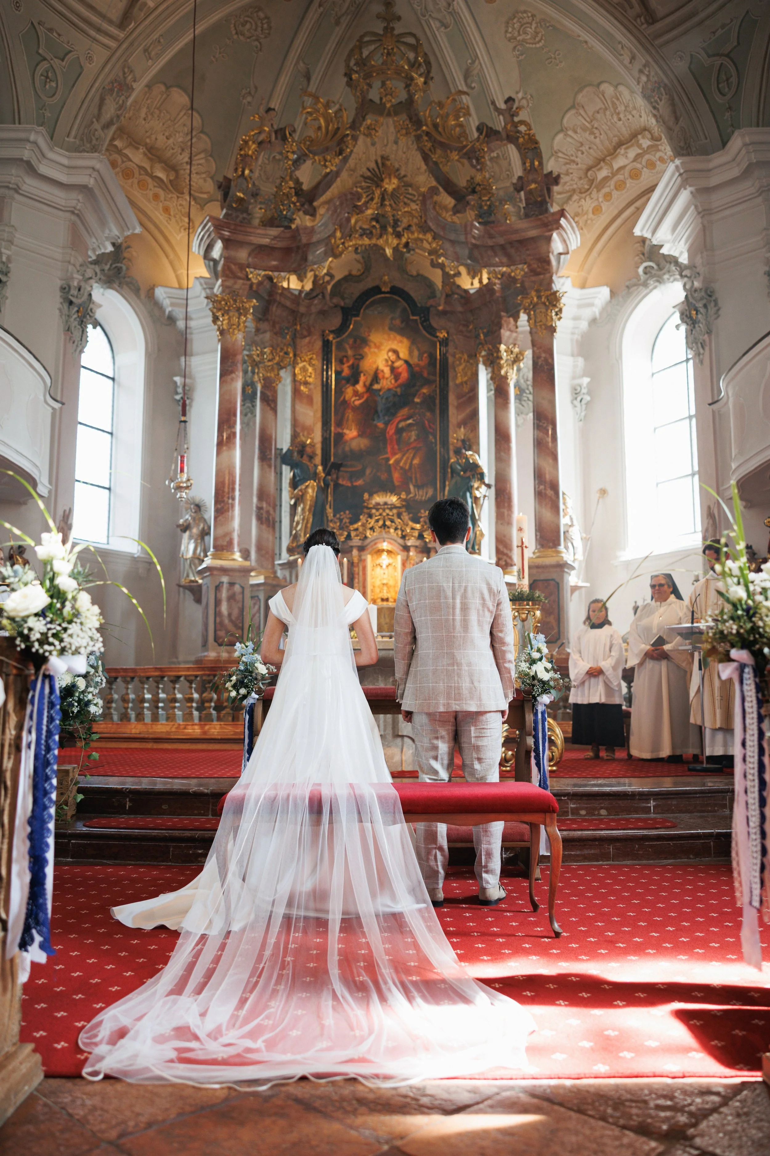Braut und Bräutigam während einer kirchlichen Hochzeitszeremonie, die vorne in einer Kirche mit barockem Altar stattfinden, mit Brautkleid, Veil und Hochzeitsanzug, Seraphinen im Hintergrund, viele Blumen und Kerzen im Raum.