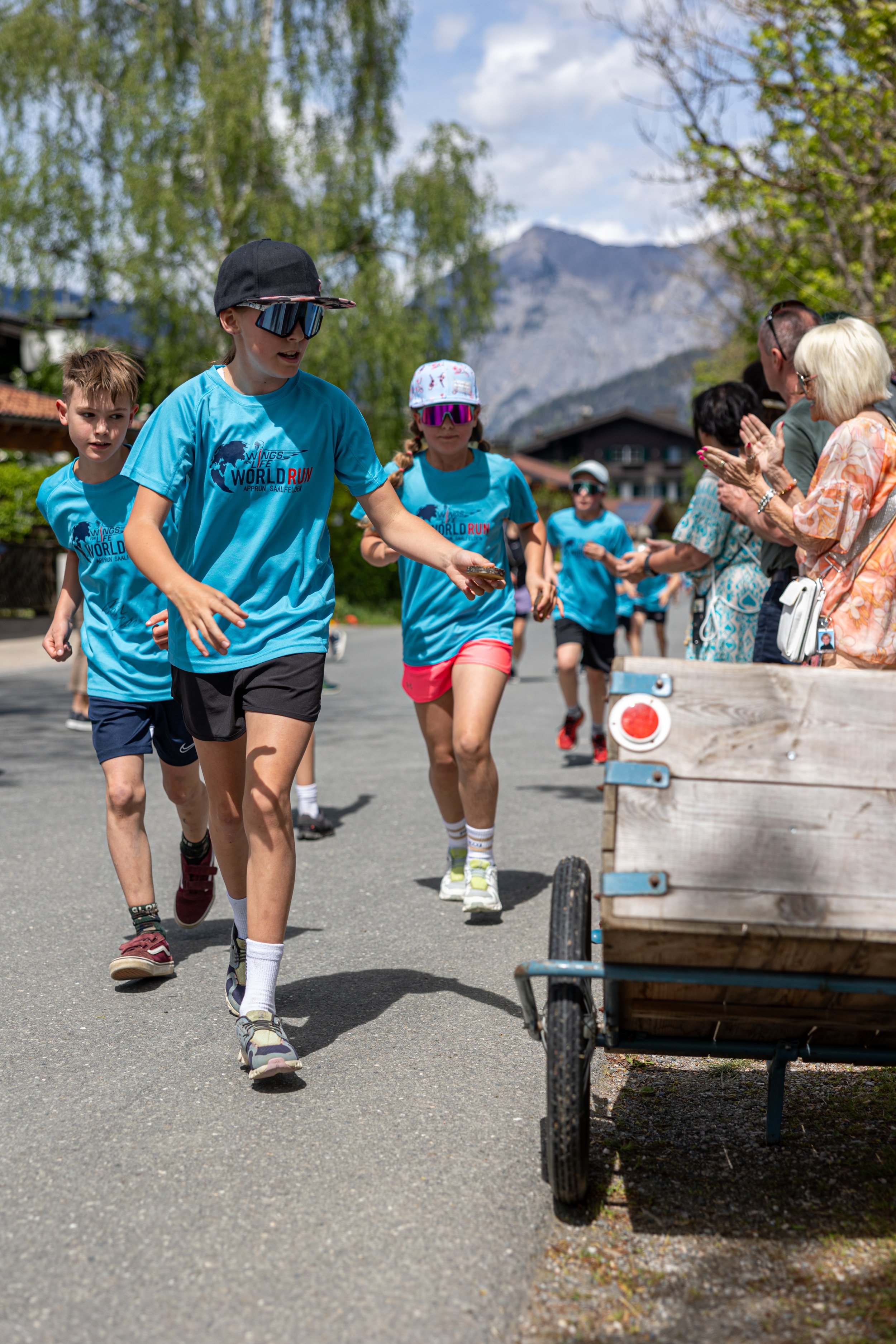 Kinder laufen bei einem Wettlauf, während Zuschauer am Spielfeldrand klatschen, im Hintergrund Berge und Bäume bei sonnigem Wetter.