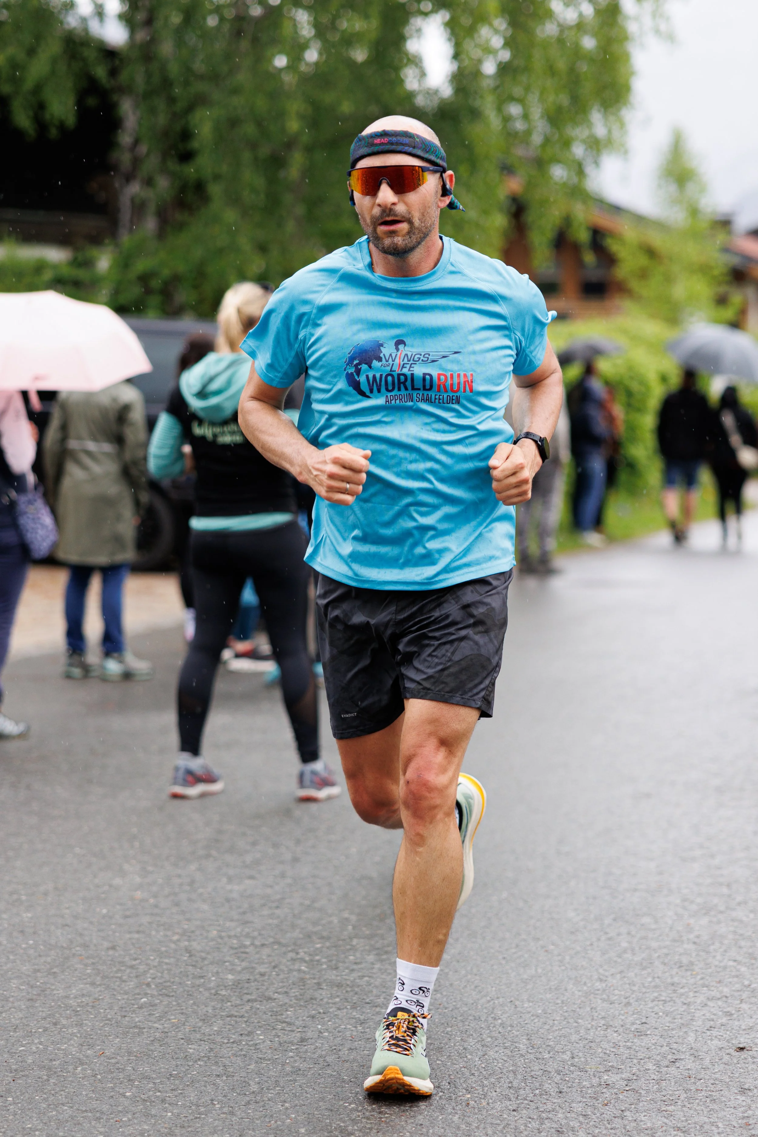 Ein männlicher Läufer während eines Straßenrennens, trägt eine blaue T-Shirt mit dem Aufdruck "WORLD RUN", schwarze Shorts, und eine Laufuhr. Im Hintergrund sind andere Läufer und Zuschauer mit Regenschirmen bei regnerischem Wetter.