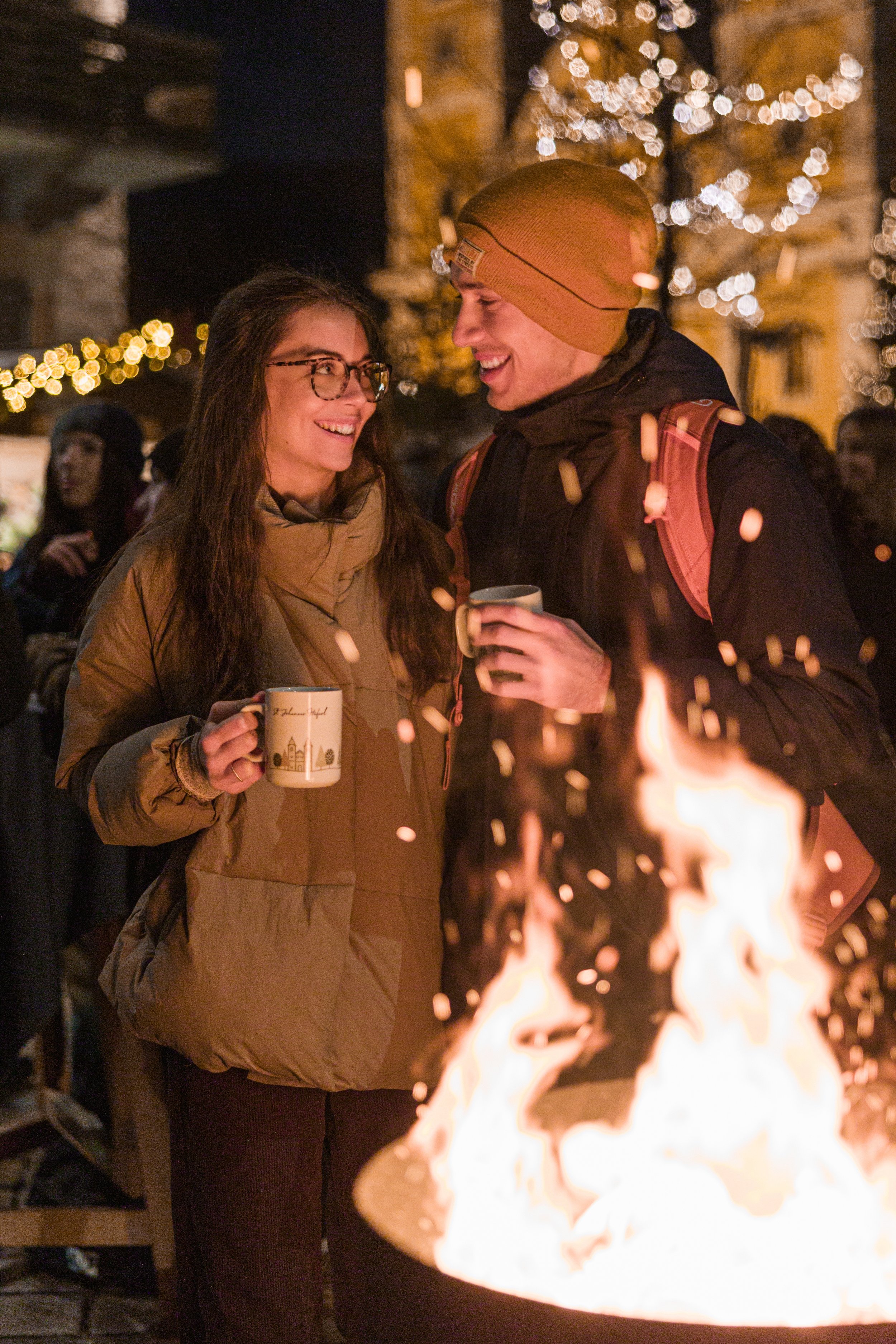 Zwei lachende Menschen, eine Frau und ein Mann, stehen bei einem Lagerfeuer, halten Tassen und genießen eine festliche Atmosphäre bei Nacht. Im Hintergrund leuchten Weihnachtslichter.