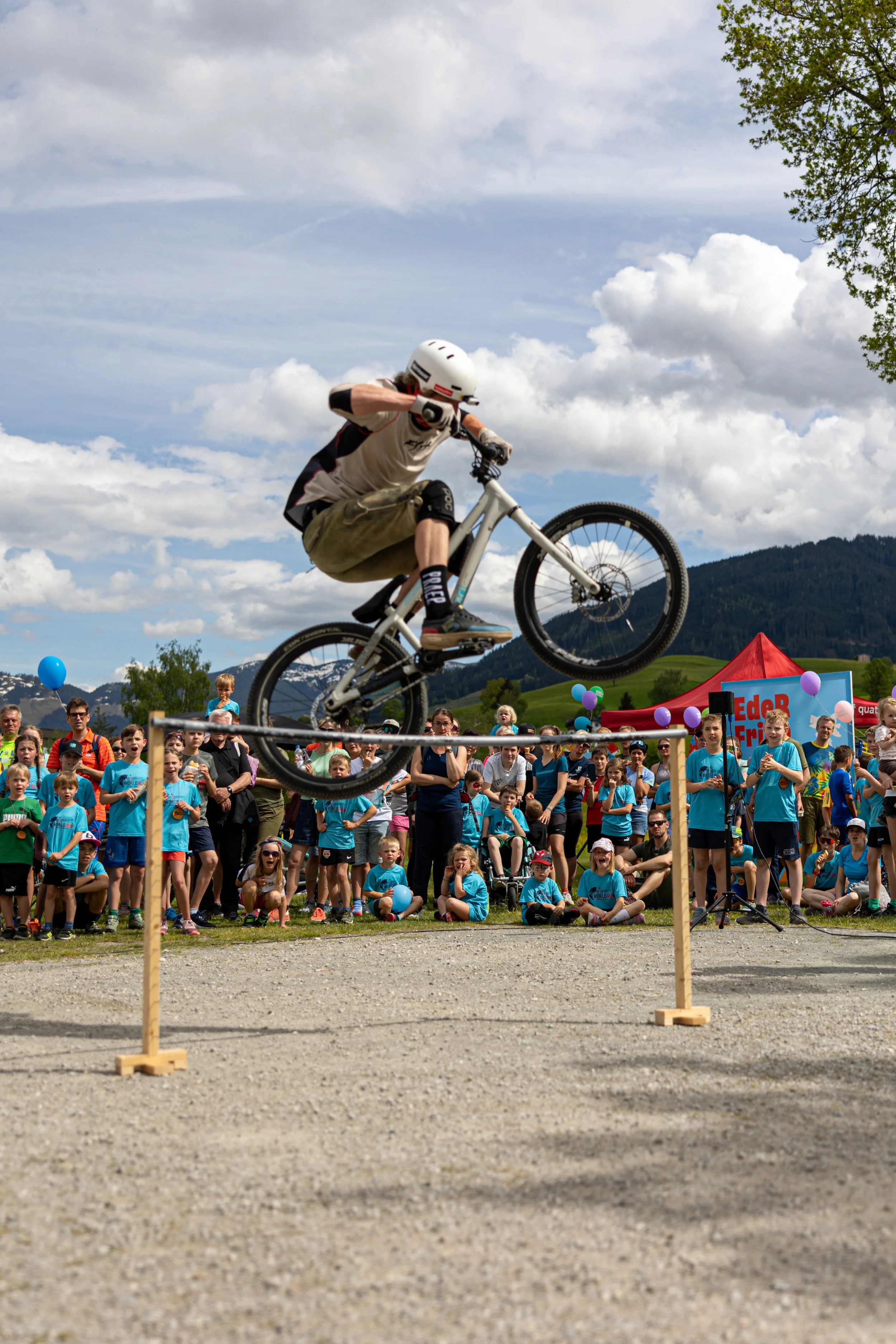 Jugendlicher fährt auf Mountainbike über eine Balken bei einem Wettbewerb, im Hintergrund steht eine große Menschenmenge und es gibt einen blauen Himmel mit Wolken.