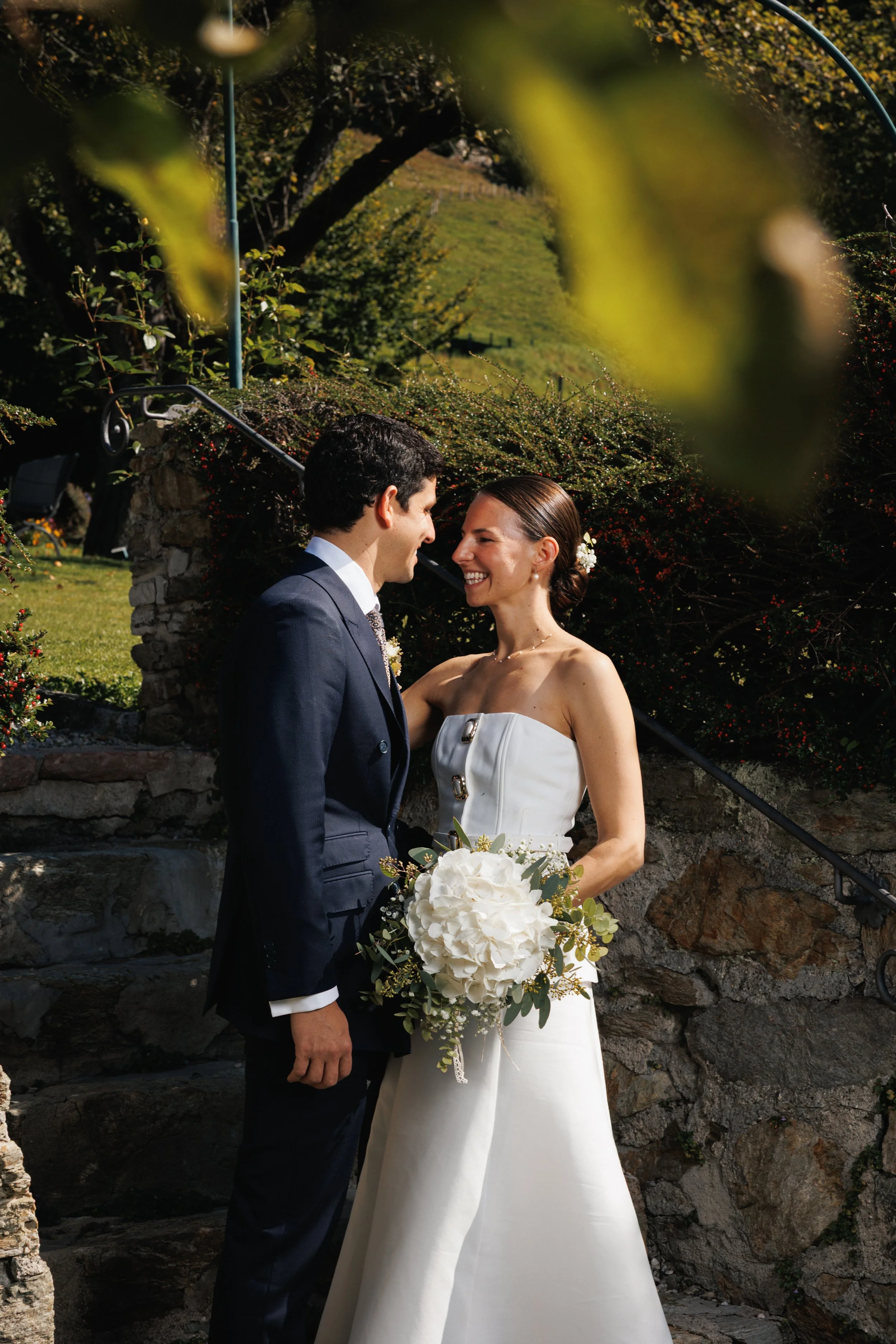 Hochzeitspaar, lachend, in einem Garten, die Braut hält einen weißen Blumenstrauß, die Szenerie ist sonnig, im Hintergrund ist Natur mit Bäumen und einem hügeligen Land.