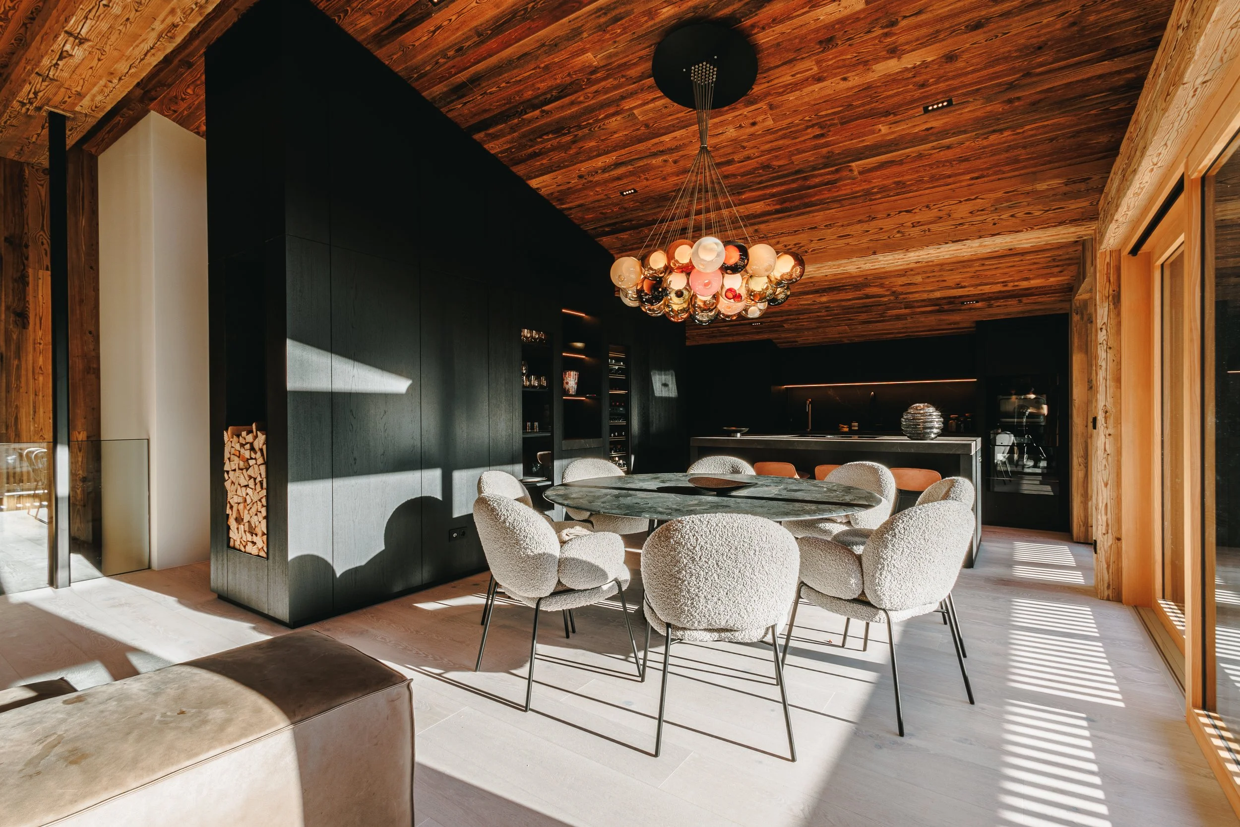 Moderne, luxuriöse Küche mit Holzdecken, einem runden Esstisch mit weißen, gepolsterten Stühlen, einer auffälligen Glaskronleuchte und großen Fenstern, die viel Tageslicht hineinlassen.