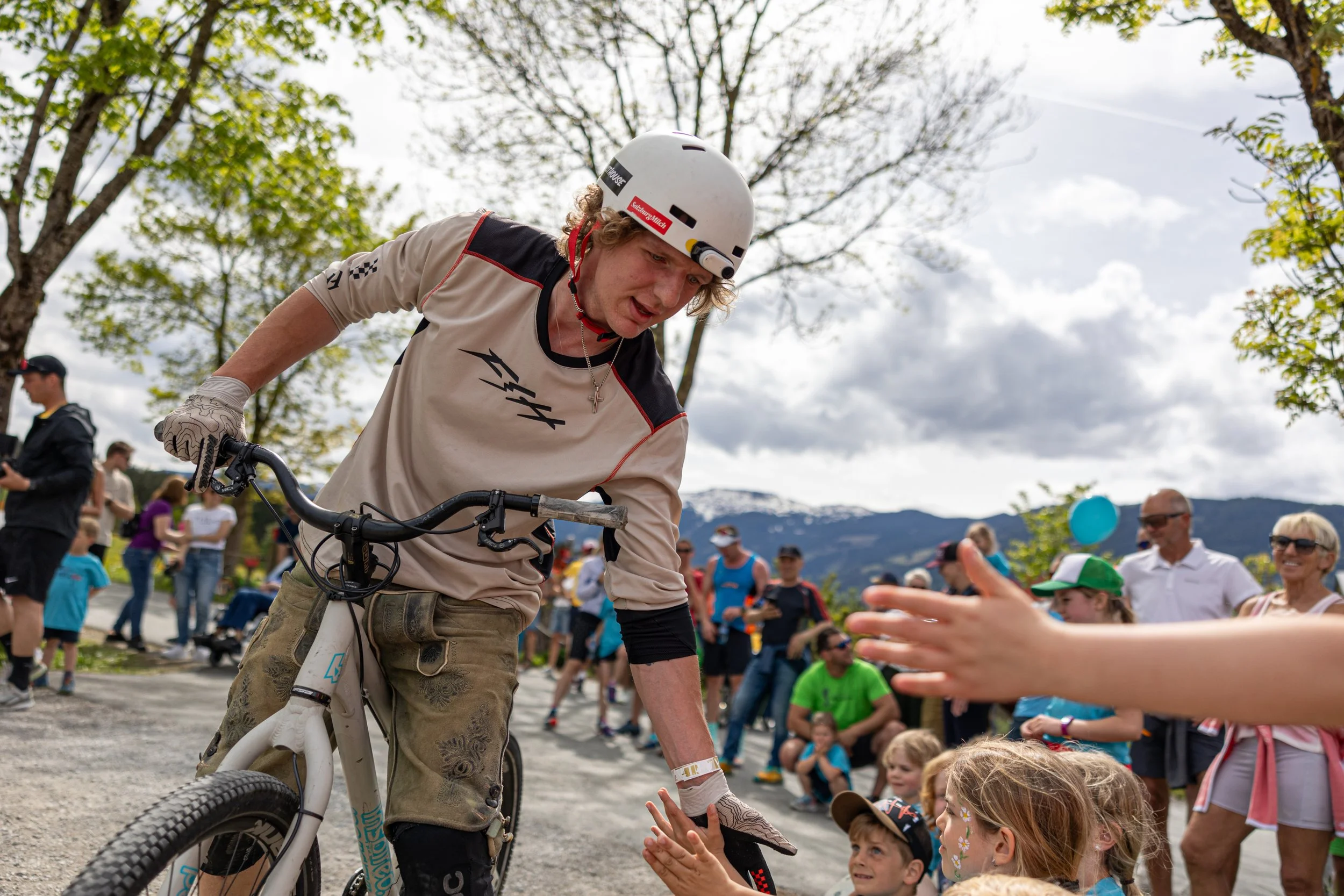 Ein junger Fahrradfahrer gibt Kinder und Erwachsenen im Freien High-Fives. Im Hintergrund sind Bäume, Berge und ein bewölkter Himmel zu sehen.