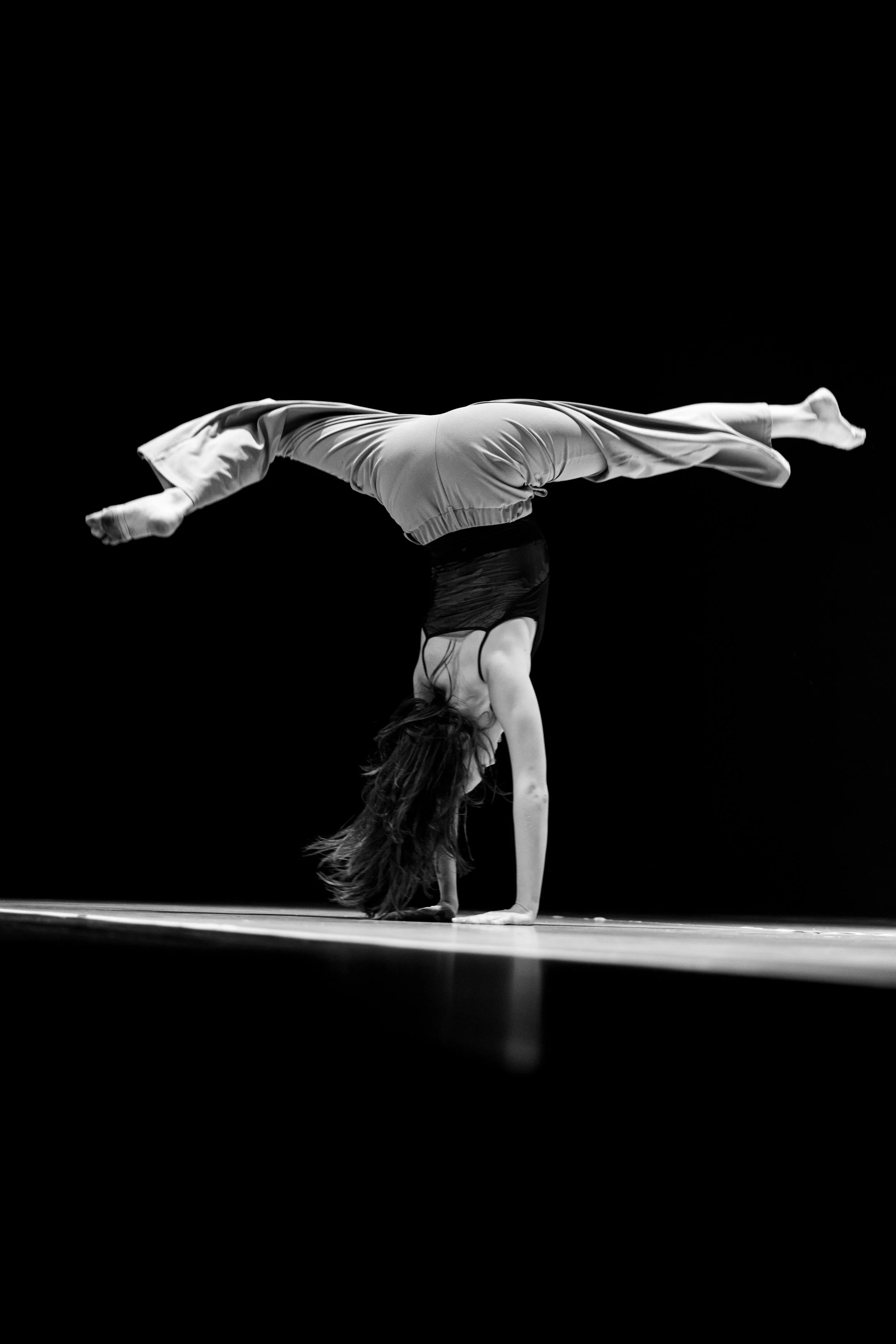 Schwarz-Weiß-Foto einer Frau in der Handstandposition, bei der sie den Körper nach unten streckt und die Beine nach oben hebt, auf einer dunklen Bühne.
