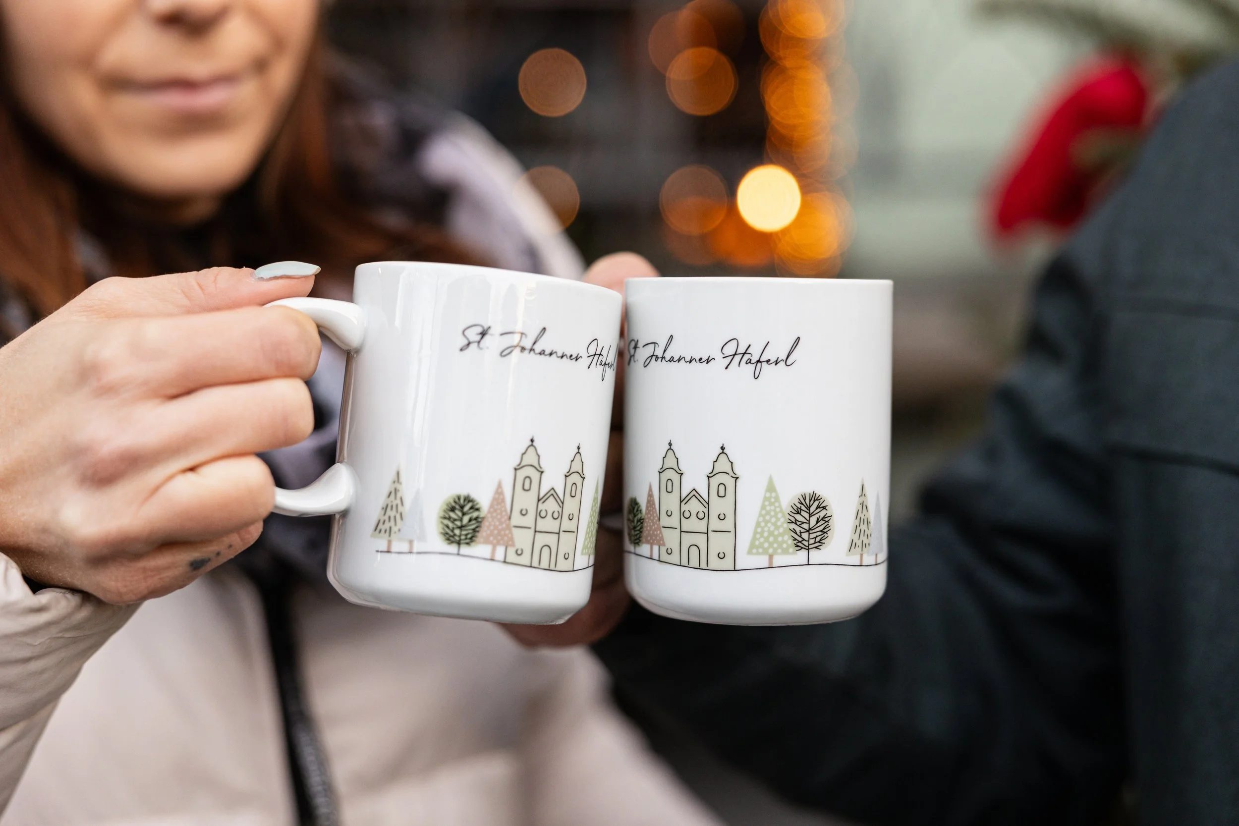 Menschen stoßen mit Tassen an, die Weihnachts- oder Wintermotive und die Aufschrift 'St. Johanner Haferl' aufweisen.