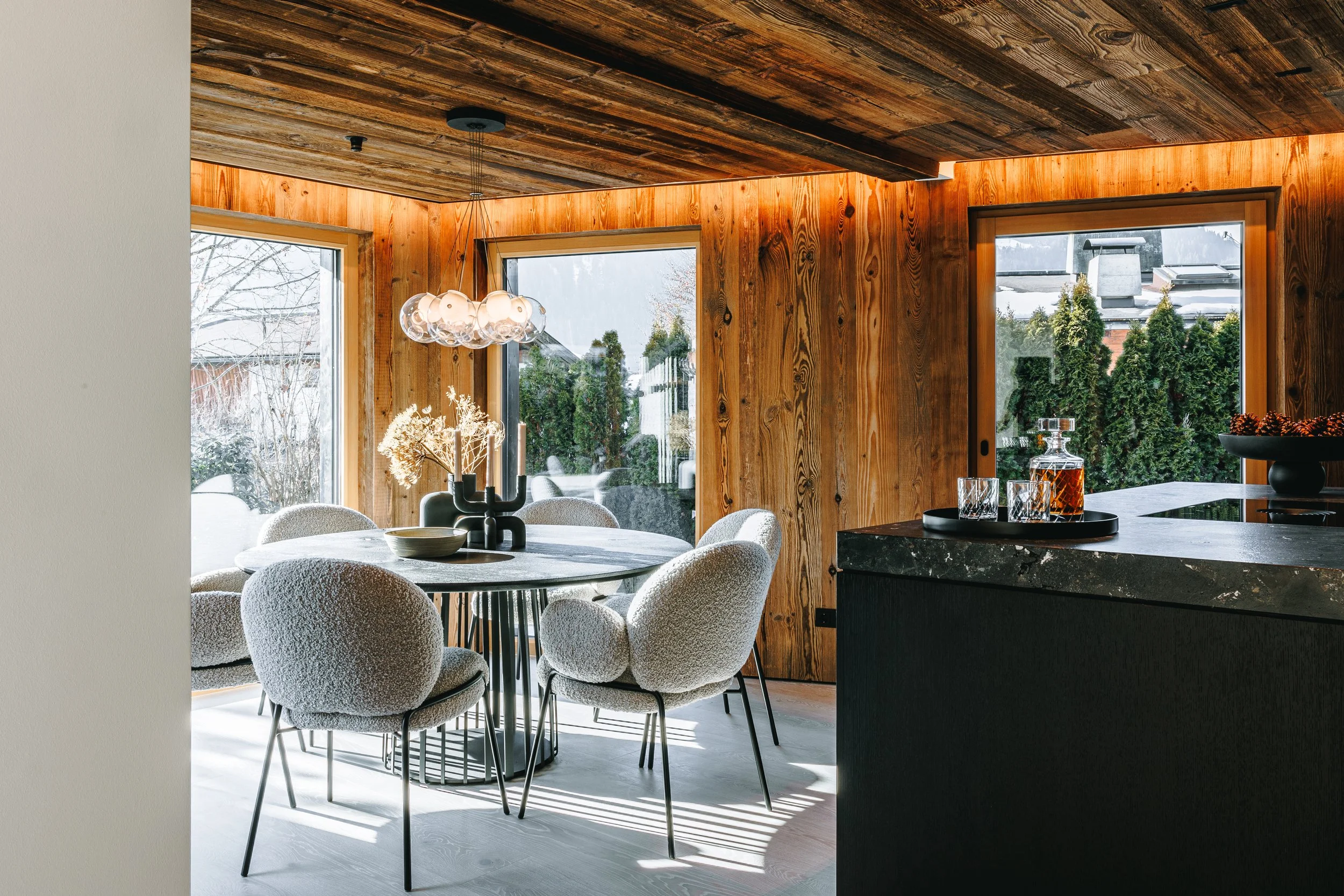 Moderner Essbereich mit runder Tisch, weißen Stühlen, holzverkleideter Wand und großen Fenstern, die Winterlandschaft zeigen.