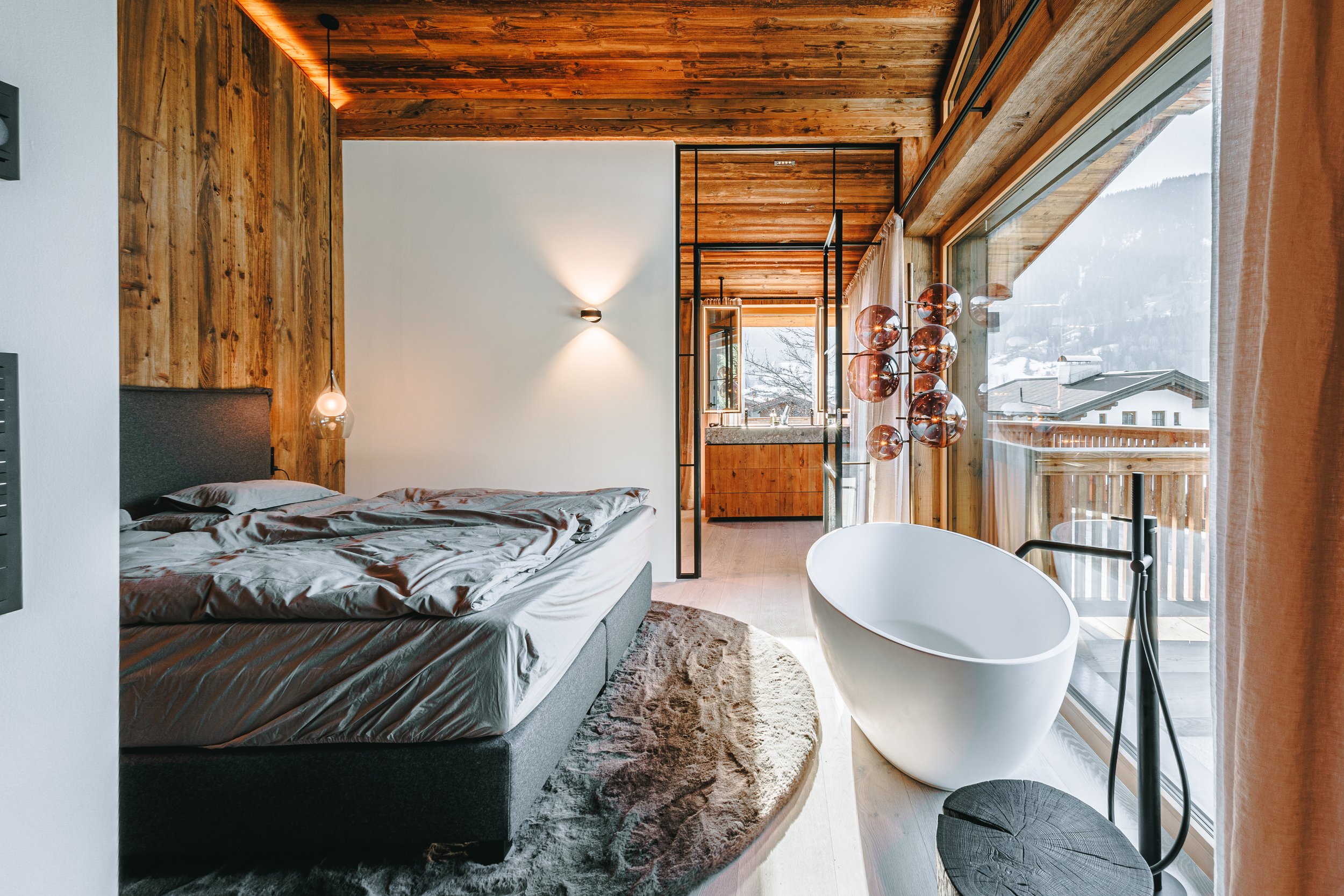 Schlafzimmer mit Holzdecke, großer Bett, freistehender Badewanne, großen Fenstern mit Blick auf Berge, moderner Beleuchtung und Dekoration.