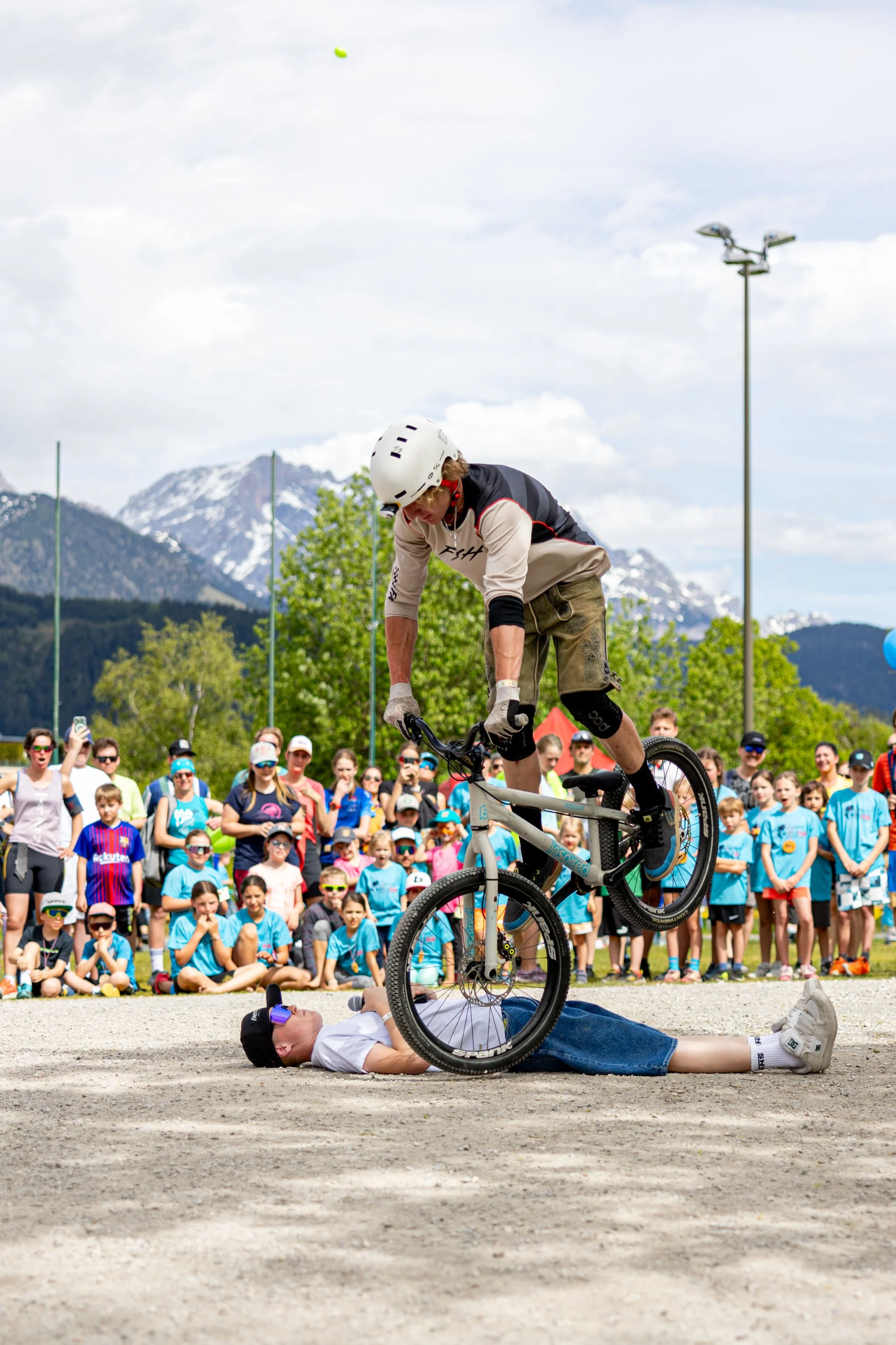 Ein Mountainbike-Fahrer springt über eine Person, die auf dem Boden liegt, während eine Menge von Zuschauern im Hintergrund zuschaut, bei einem Outdoor-Event mit Bergen im Hintergrund.