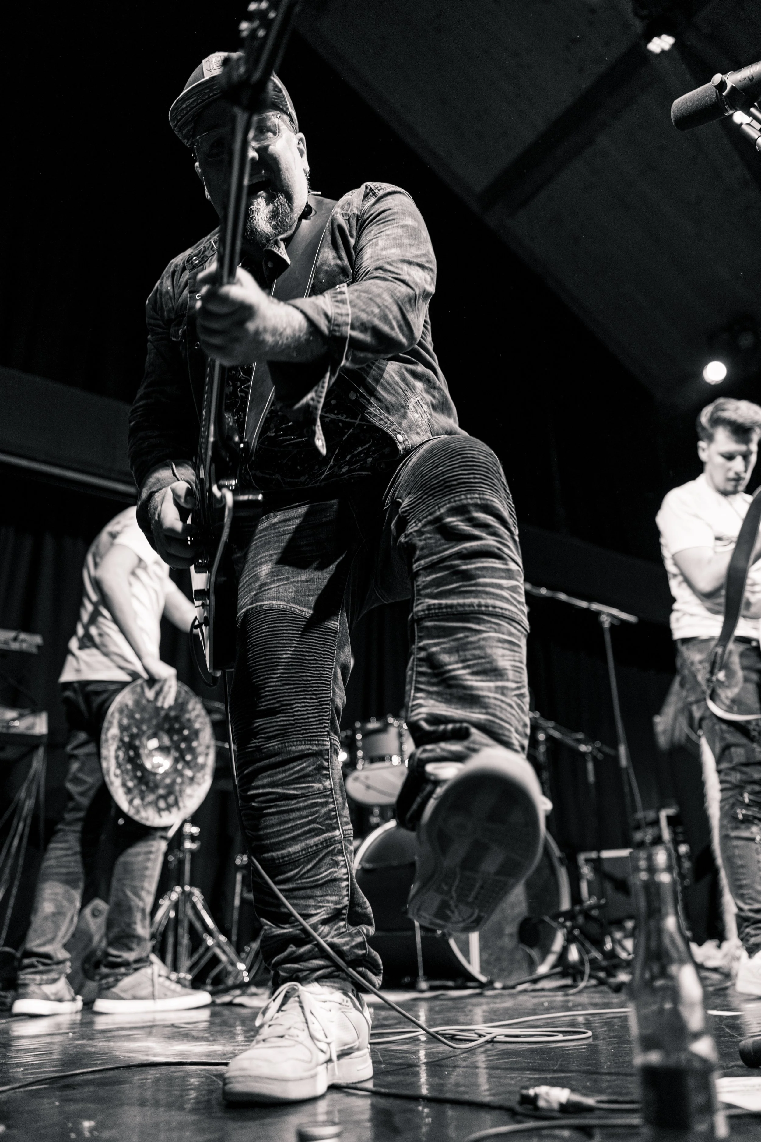 Ein Musiker während eines Auftritts, spielend Gitarre, im schwarzen und weißen Foto. Andere Bandmitglieder sind im Hintergrund zu sehen.