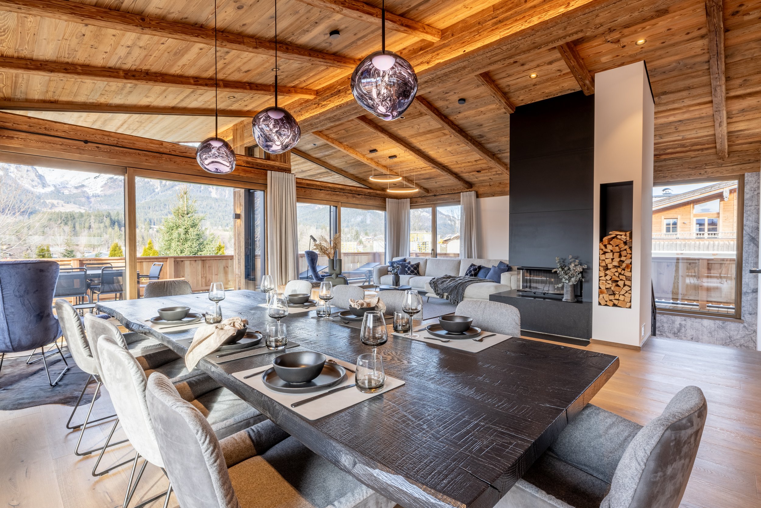 Offenes Wohnzimmer und Esszimmer in einem Chalet, mit Holzdecke, großen Fenstern und modernem Dekor, Blick auf Berge im Hintergrund.