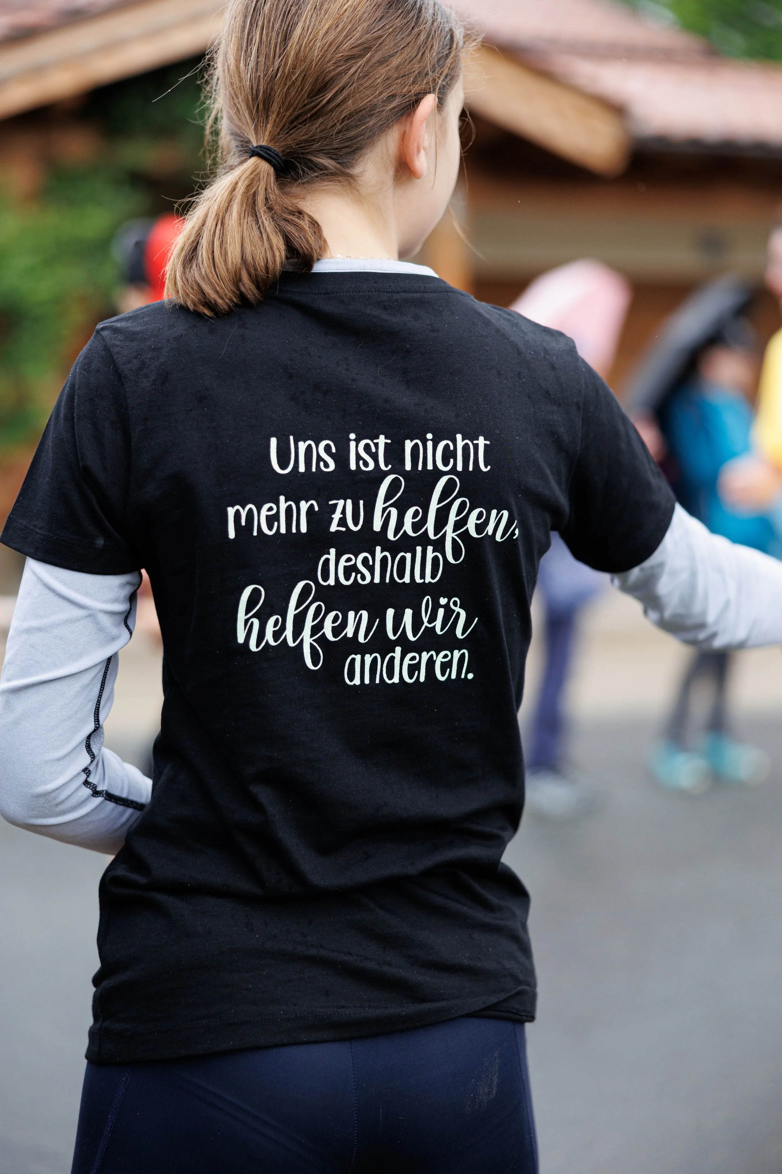 Rückansicht einer Frau mit braunen Haaren in einem Pferdeschwanz, die einen schwarzen T-Shirt trägt. Auf dem T-Shirt steht auf Deutsch: "Uns ist nicht mehr zu helfen, deshalb helfen wir anderen."