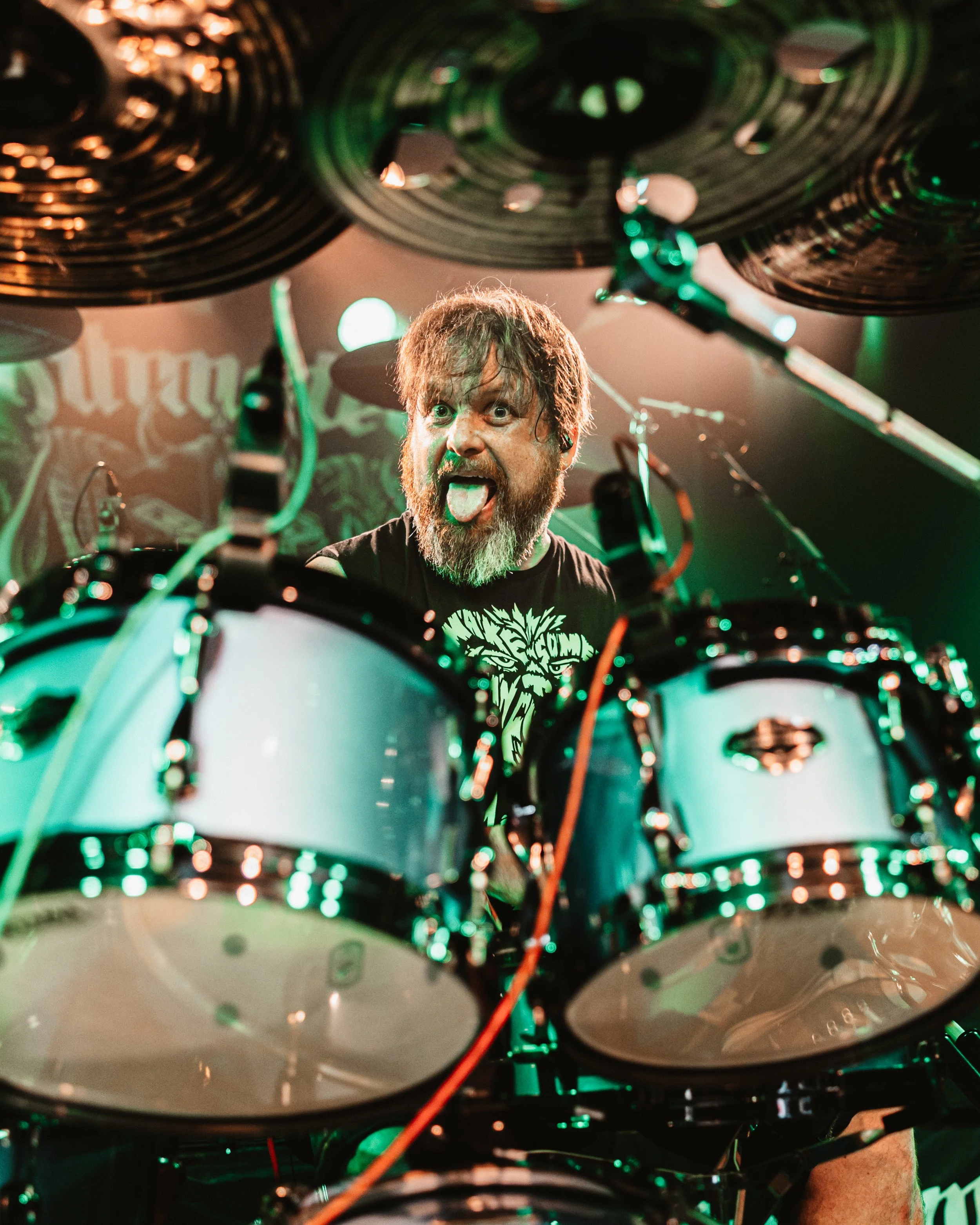 Musiker hinter einem Schlagzeug, der die Zunge herausstreckt, auf einer Bühne mit grüner Beleuchtung.