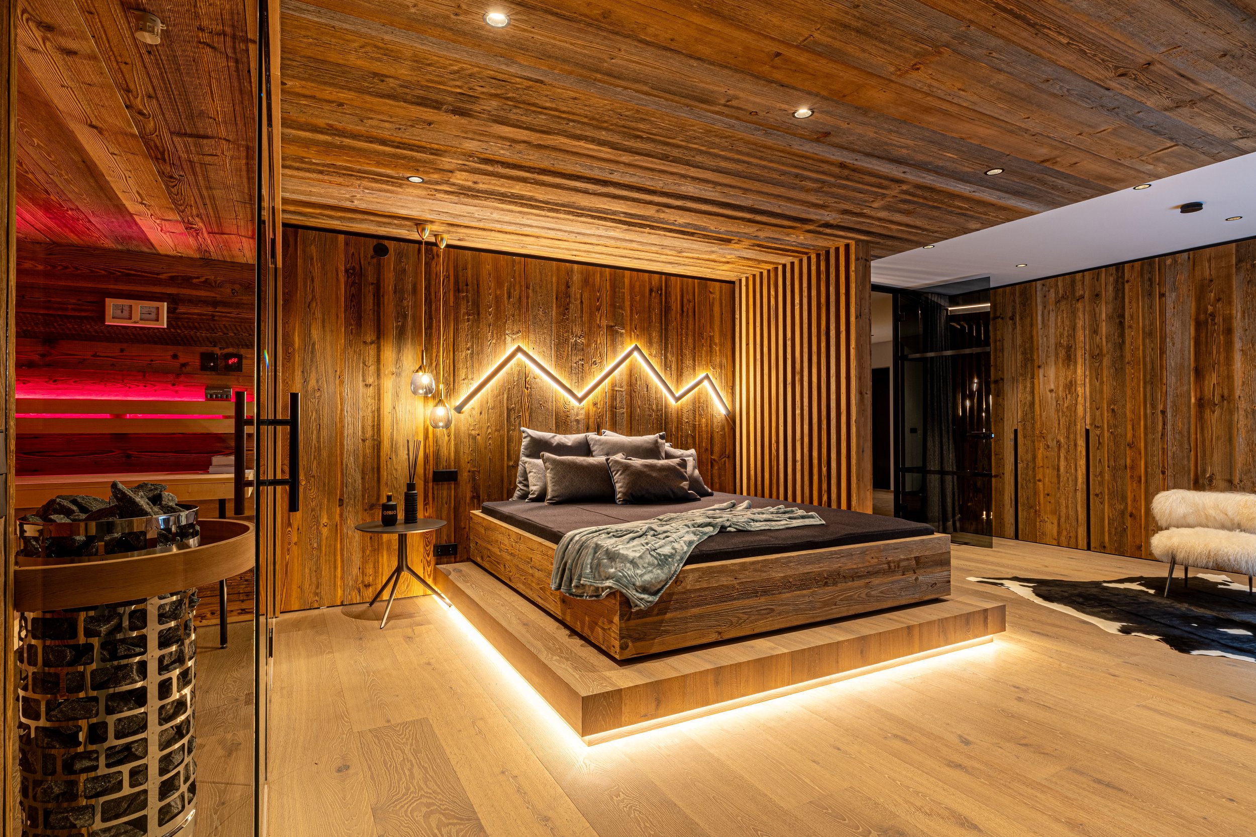 Schlafzimmer mit Holzdecke und -wänden, Bett mit schwarzen und grauen Kissen, moderne Beleuchtung und Dekoration, Holzsitz mit Sauna im Hintergrund, kuscheliges weißes Sitzmöbel, Tierfell auf Teppich.