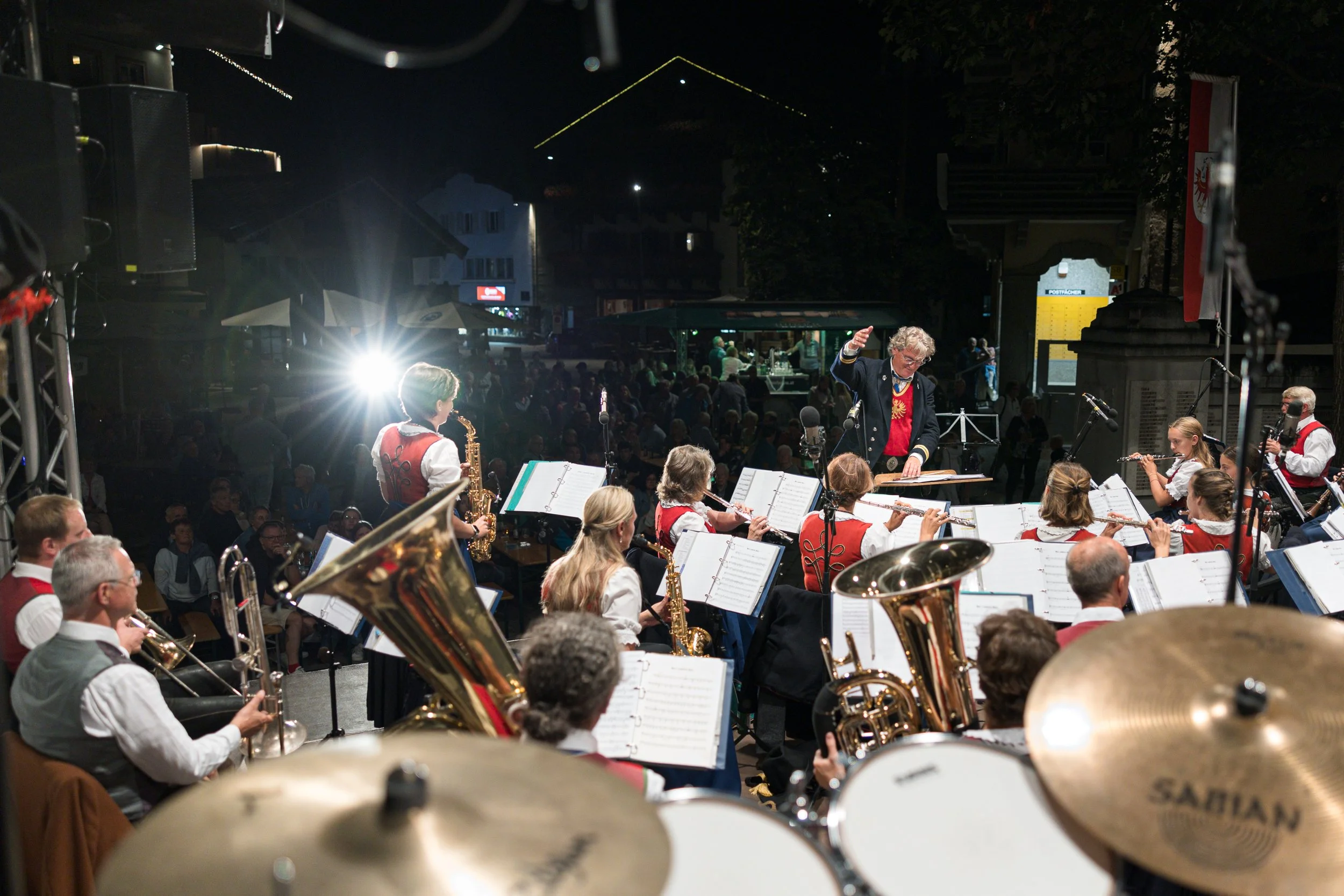 Orchestermusiker spielen bei einem Konzert im Freien, im Hintergrund ein Publikum und Stadtgebäude bei Nacht.