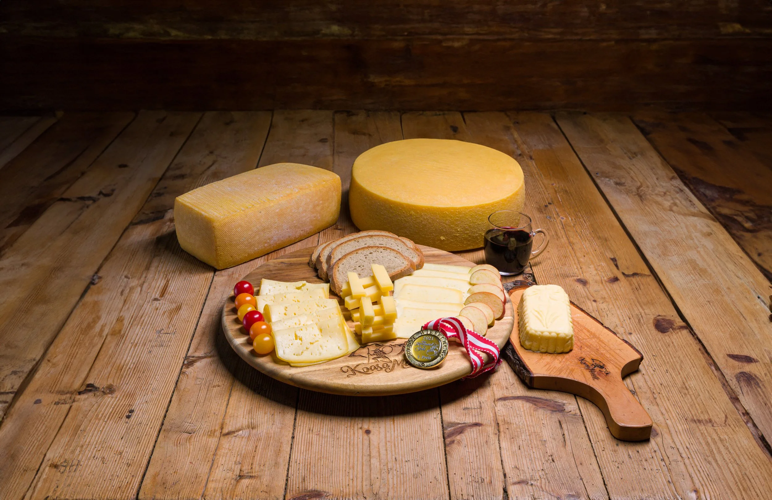 Brett mit Käsesorten, Tomaten, Wurst, Wein und Brot, daneben zwei große Käselaibe auf Holz unter Sitz im rustikalen Holzraum.