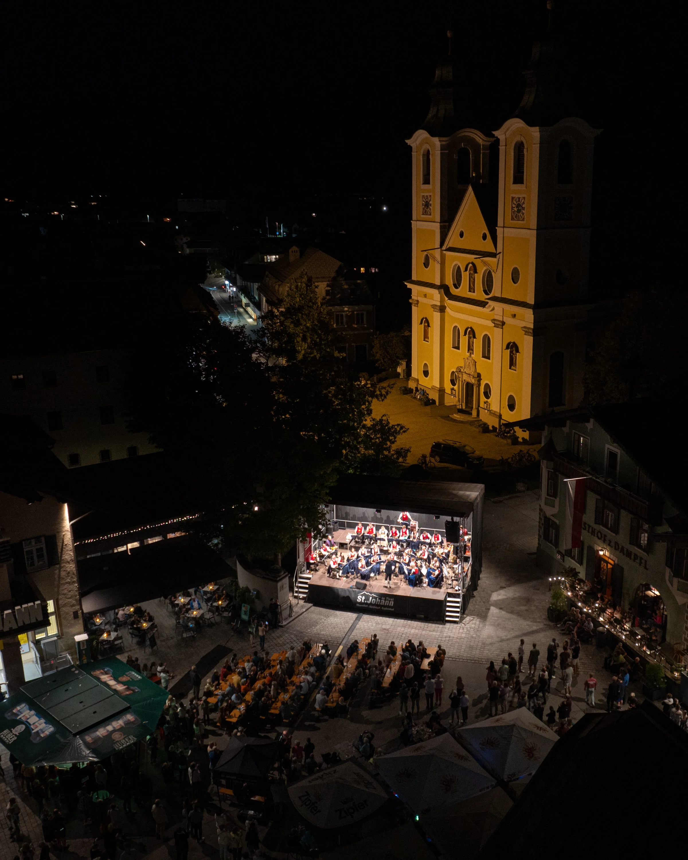 Nachtaufnahme eines festlichen Open-Air-Orchesterkonzerts vor einer barockaden Kirche in einer kleinen Stadt, mit Zuschauer auf Sitzgelegenheiten und Tischen im Freien.