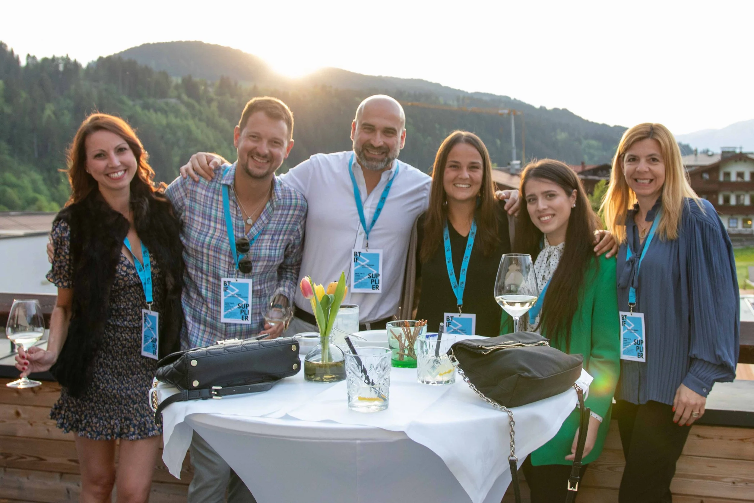 Eine Gruppe von sieben Menschen beim gesellschaftlichen Treffen auf einer Terrasse, im Hintergrund Berge und Bäume bei Sonnenuntergang, sie tragen Namensschilder, einige halten Weingläser, einen Tisch mit Dekoration und Getränken davor.
