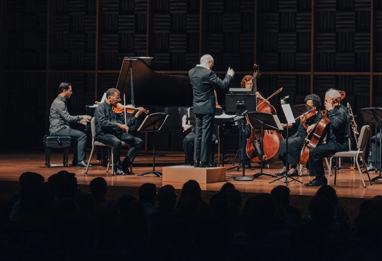 Chamber orchestra performing on stage with a conductor, violinist, pianist, cellists, and a bassist.