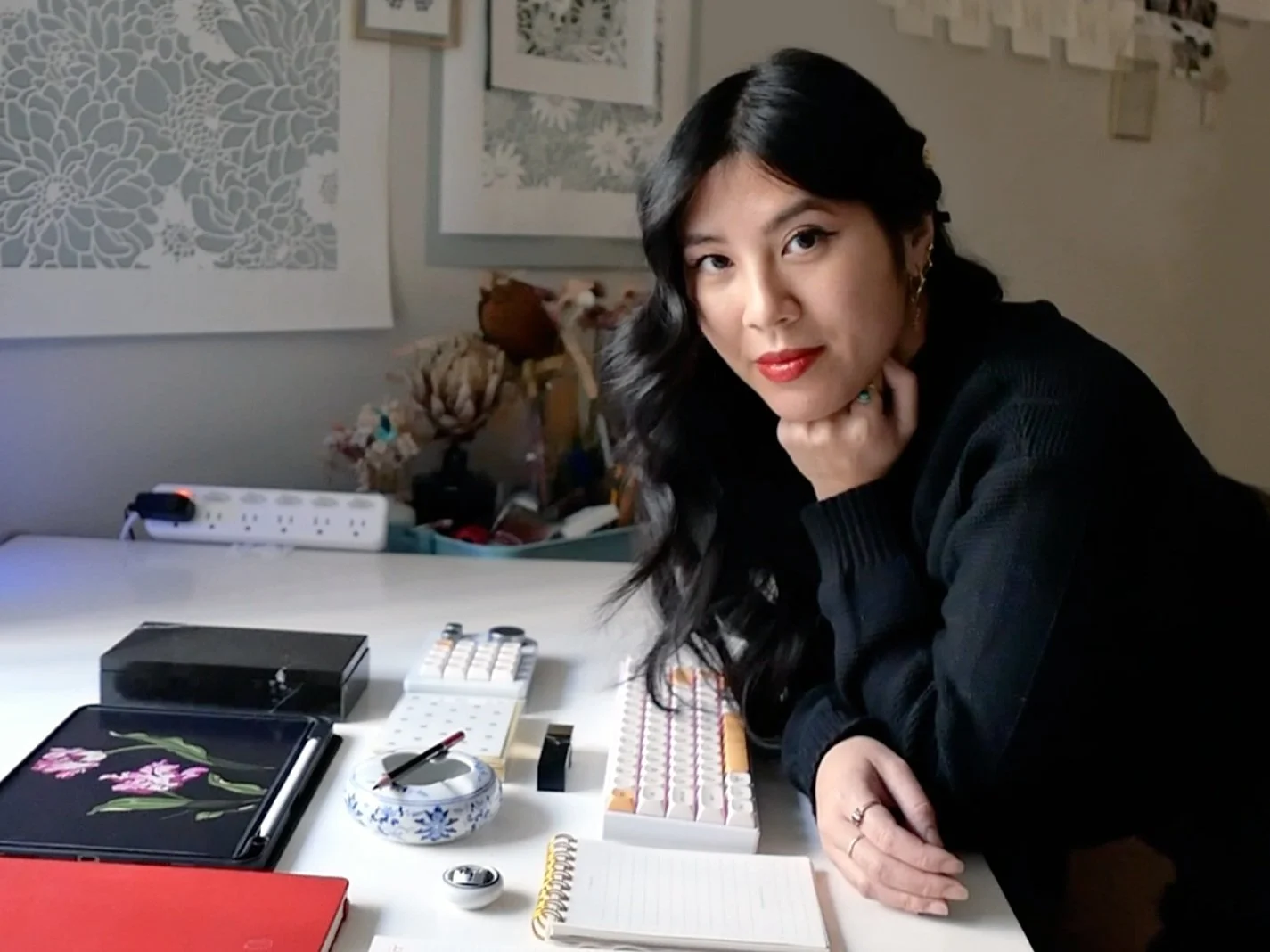 A woman with long black curly hair and red lipstick leaning on a white desk with a notebook, a tablet with floral design, a small white calculator, and various office supplies. The background includes framed artwork and a lamp.