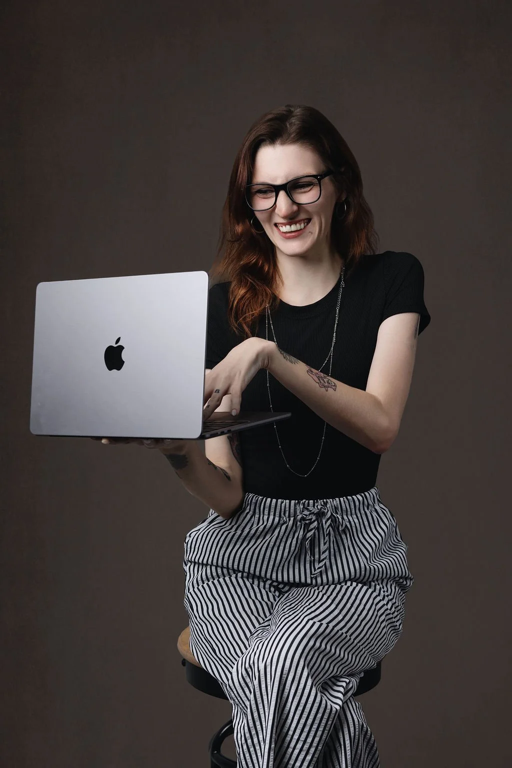 A woman with glasses, tattoos, and a black shirt sitting on a stool, smiling and looking at a silver MacBook. Kylee Ellis, Client Concierge at Monroe & Co Boudoir