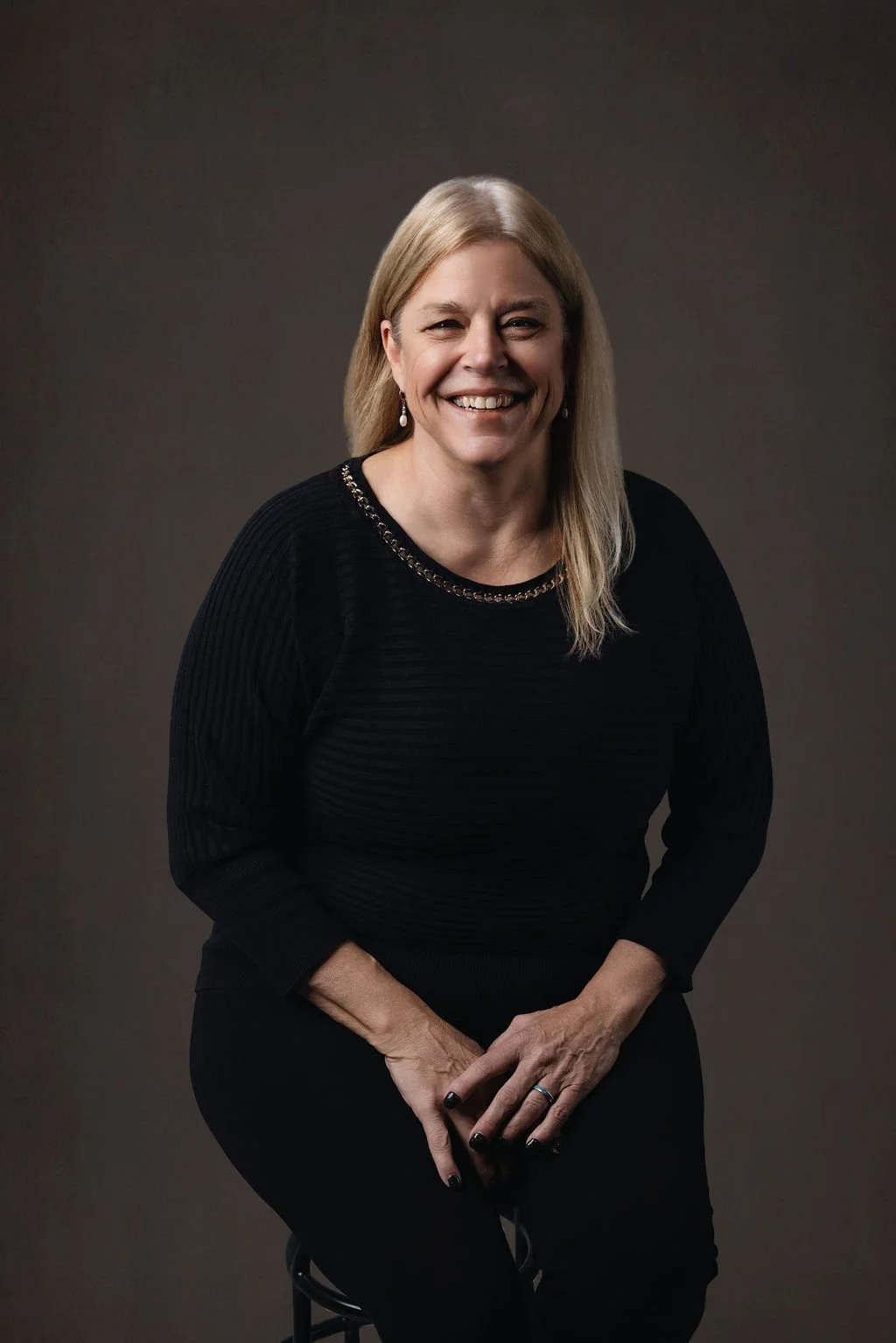 A woman with blonde hair smiling, sitting against a plain dark background, wearing a black top with a chain detail around the neckline. Eileen Ellars, Studio Director at Monroe & Co Boudoir