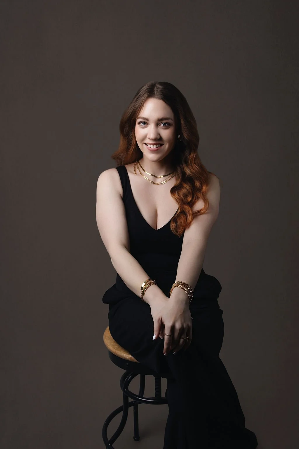 A woman with straight brown hair, fair skin, wearing a black polka-dot blouse, gold jewelry, and posing with her arms crossed, against a dark background.