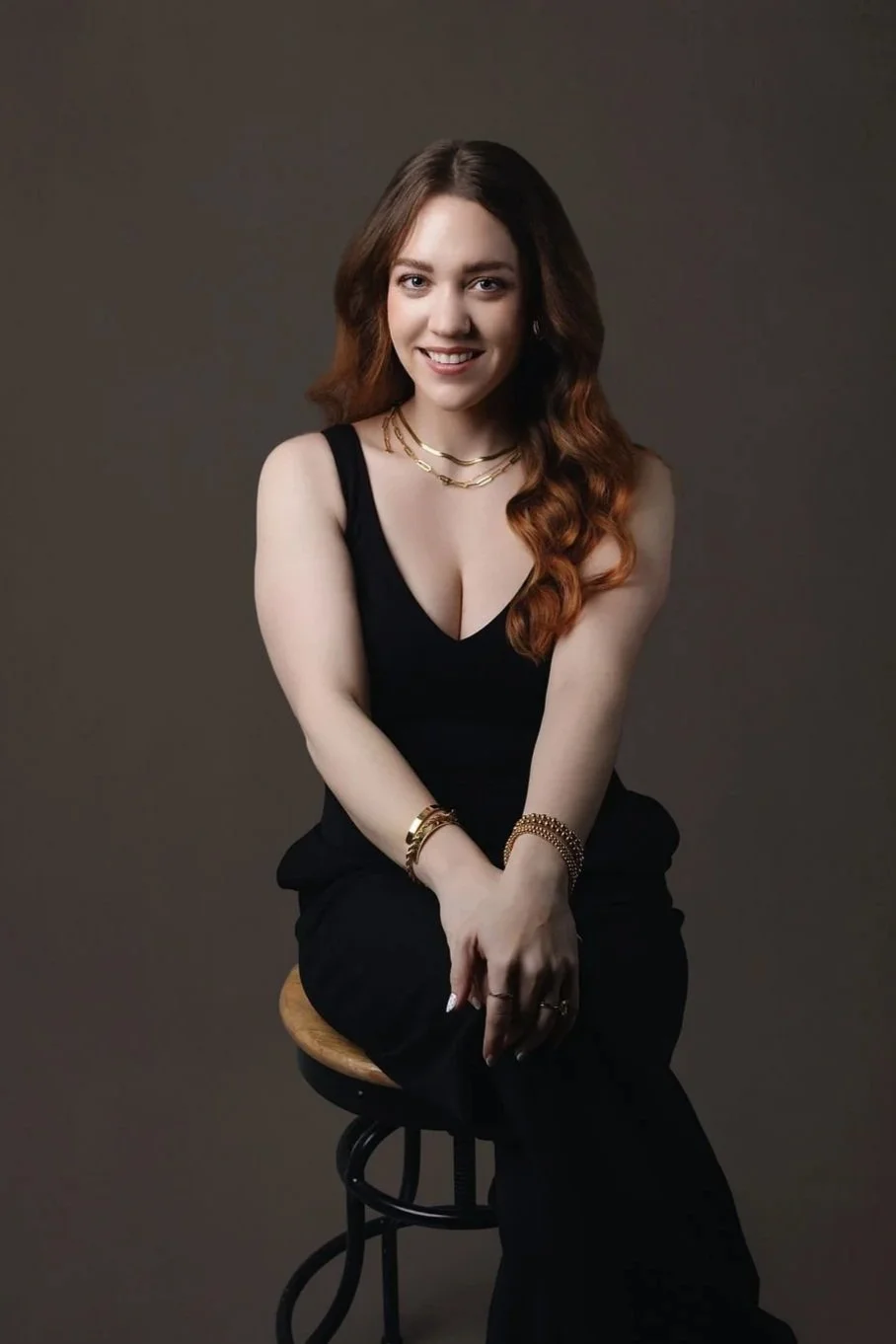 A woman with straight brown hair, fair skin, wearing a black polka-dot blouse, gold jewelry, and posing with her arms crossed, against a dark background.