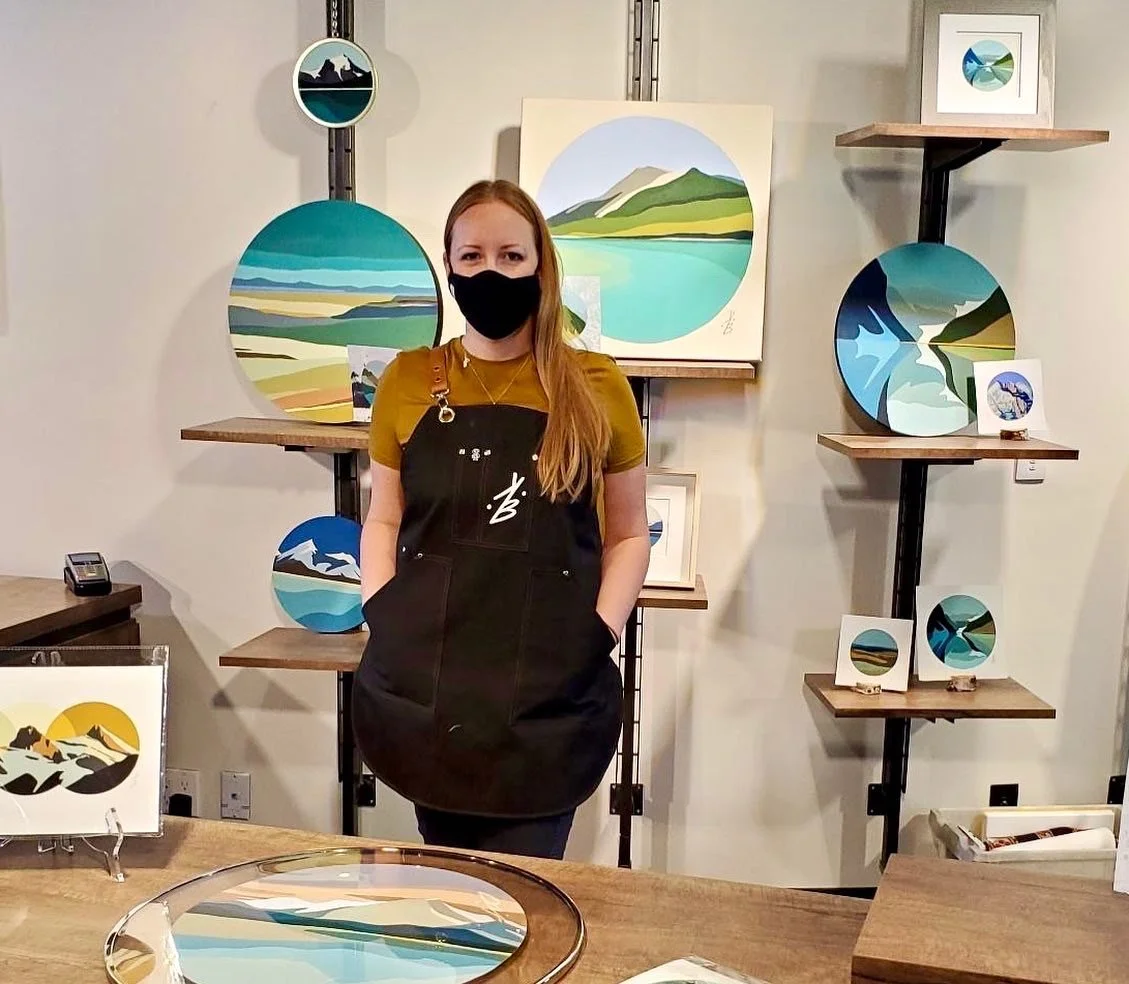 A woman wearing a black mask and apron standing in front of a display of landscape paintings featuring mountains and water in various circular arrangements.