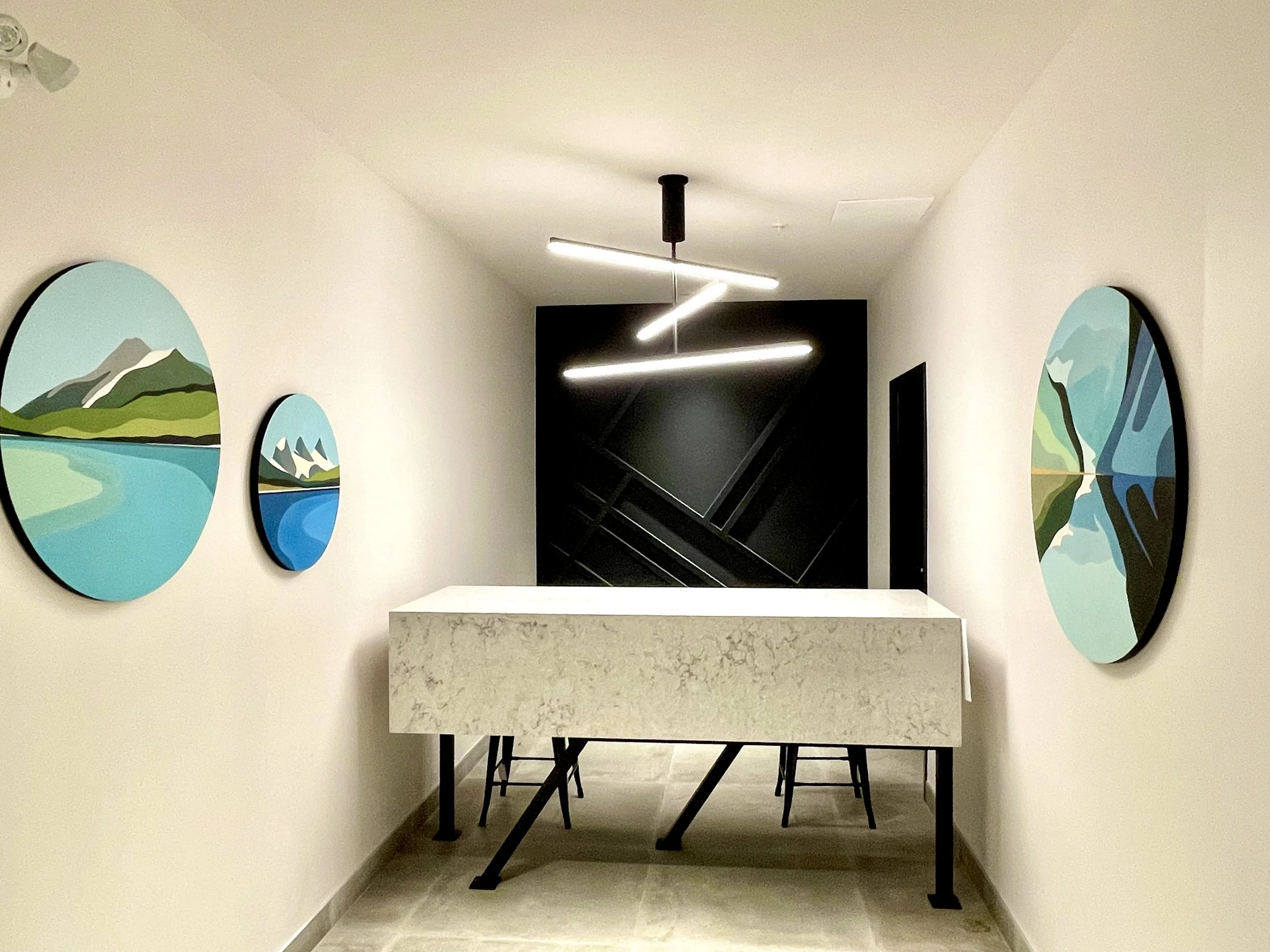 Modern minimalist hallway with white walls decorated with landscape paintings, a rectangular stone table, and black chairs, with an abstract light fixture on the ceiling.