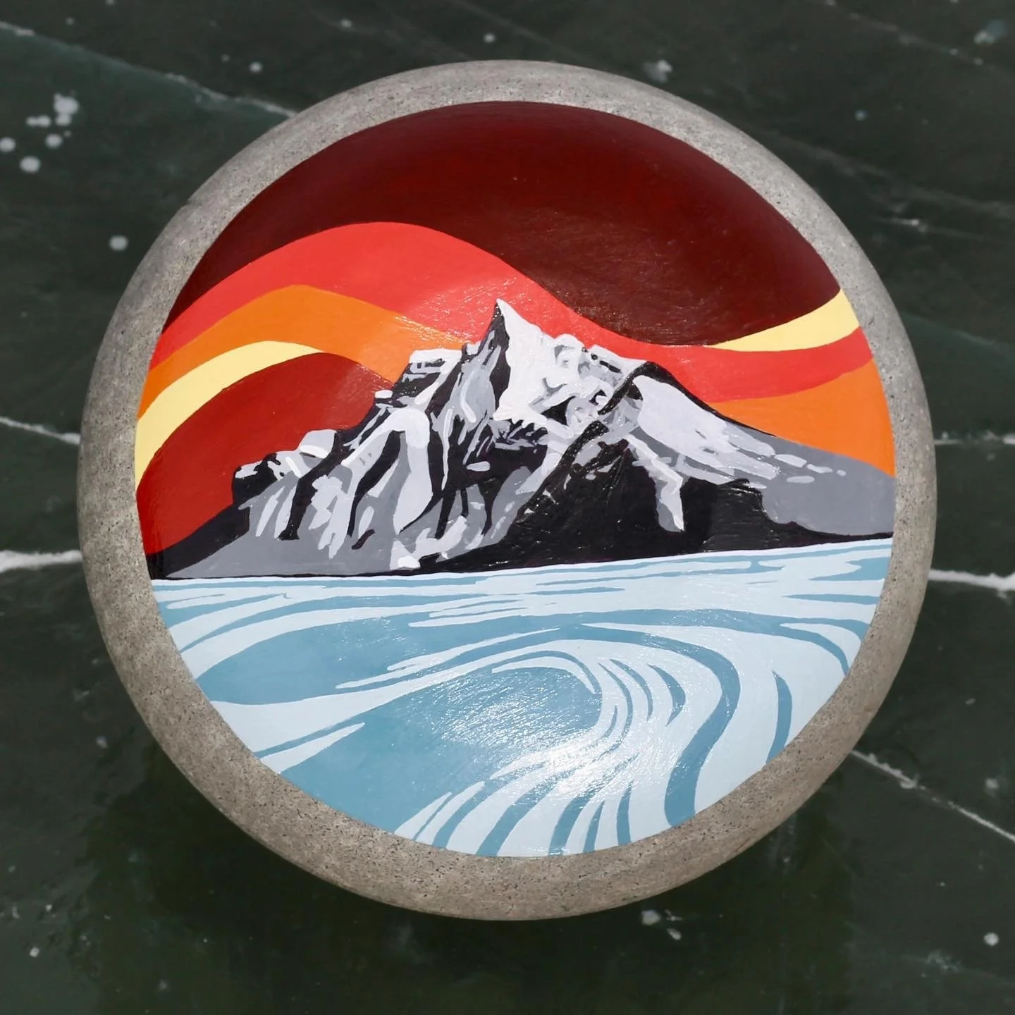 Painted rock with a mountain landscape, snow-capped peaks, colorful sunset sky, and icy water, surrounded by a gray concrete border.