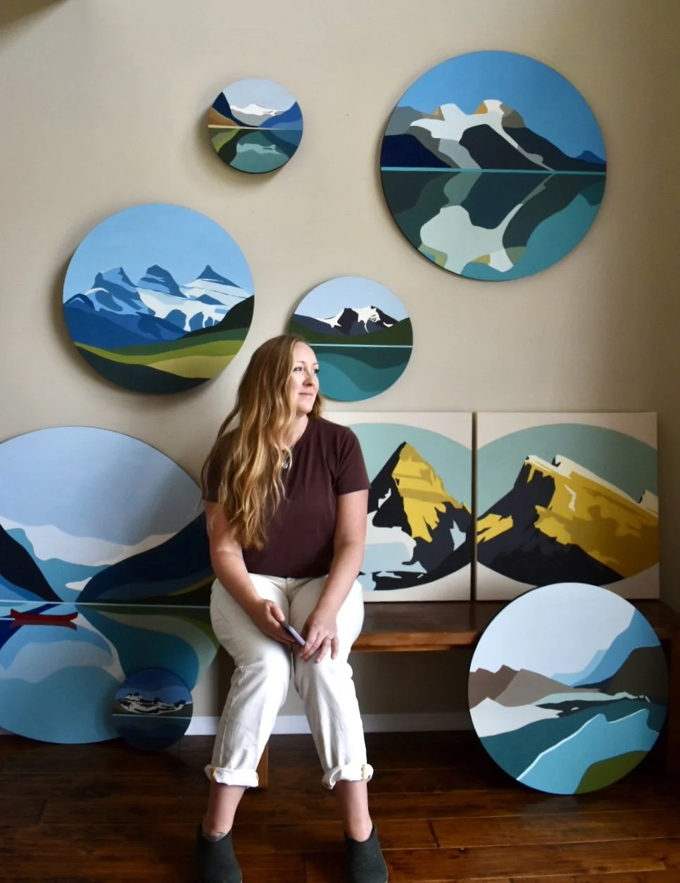 Woman sitting in front of a wall decorated with circular landscape paintings of mountains, lakes, and nature scenes.