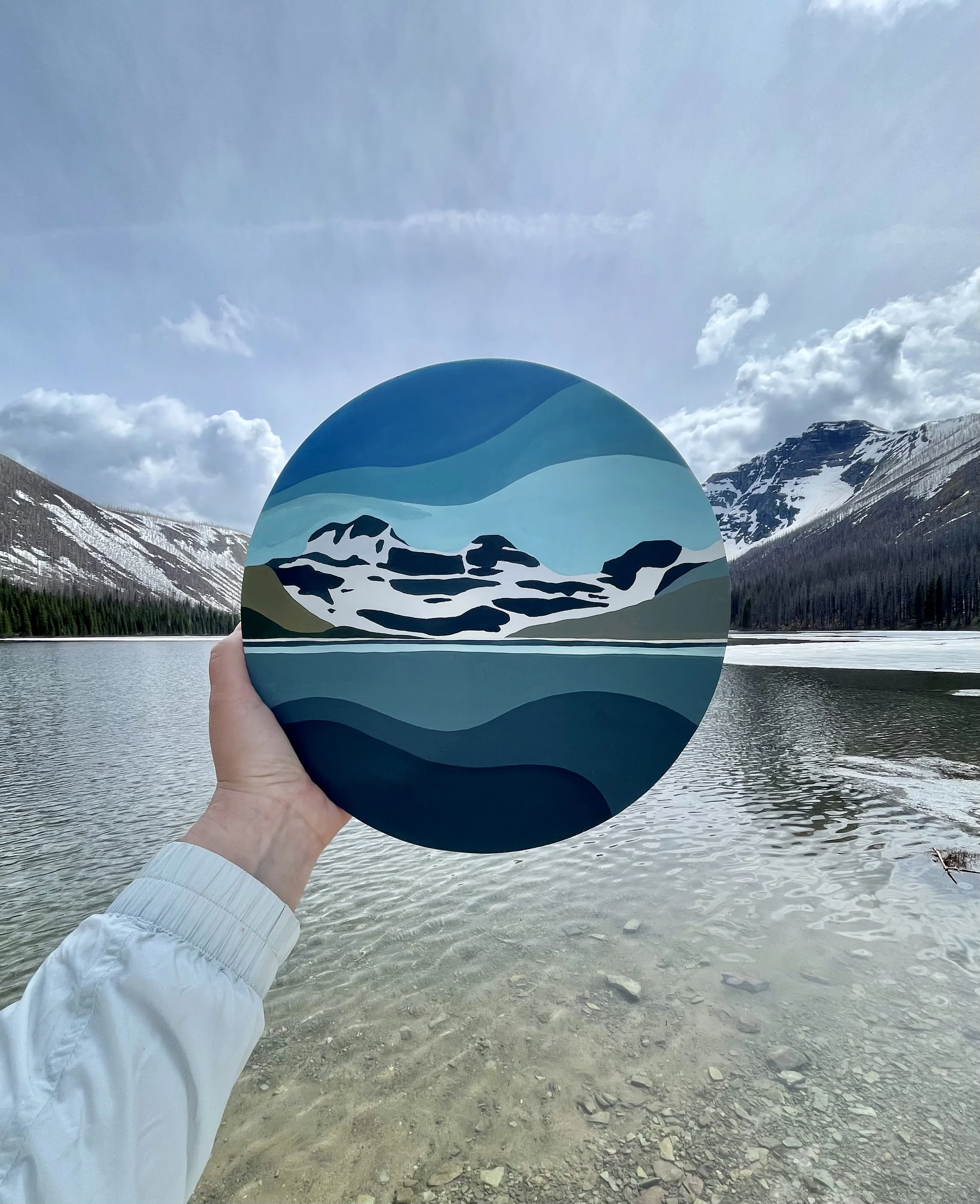 A hand holding a circular painting of mountains and water, with a mountain lake landscape in the background.