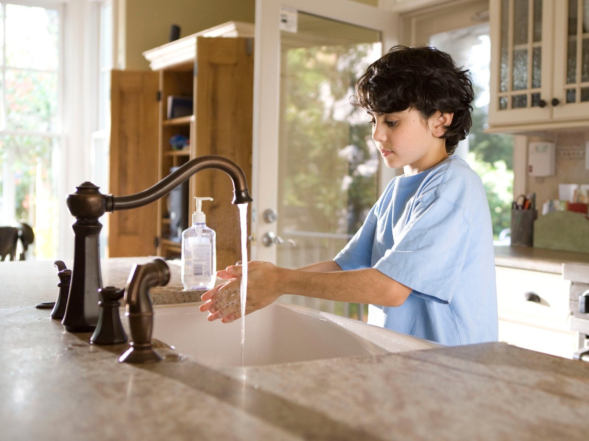 A child in a blue shirt washing their hands with soap and water at a kitchen faucet