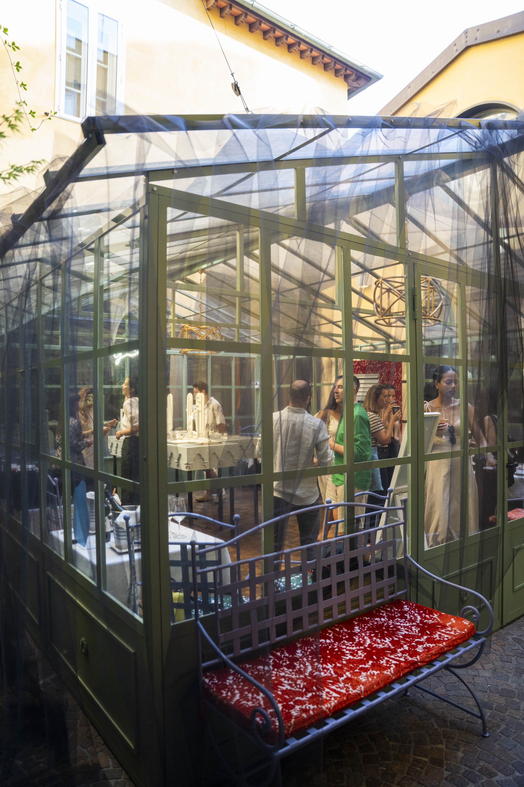 A glass greenhouse with people inside, surrounded by a green metal frame, with various tables set for a gathering, and a red and white bench outside.