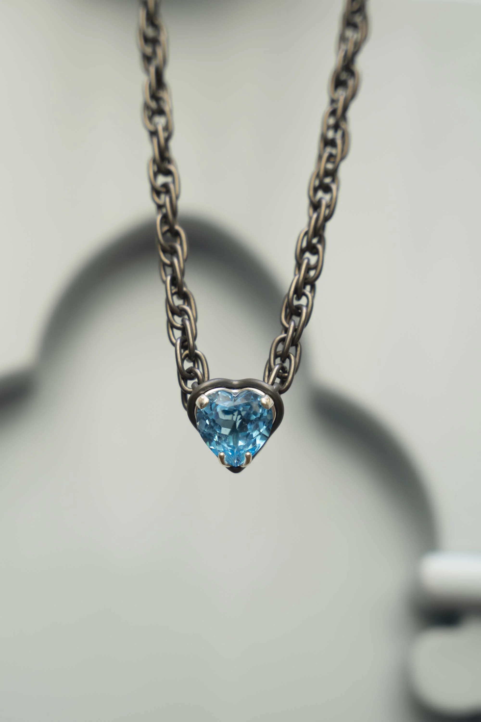 Gi by Giselle Open Heart Necklace, 13-carat blue topaz heart pendant on titanium chain, hanging against a wall