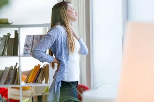 Woman standing in her living room with one hand on her back and one hand near her neck with her head tilted in discomfort.